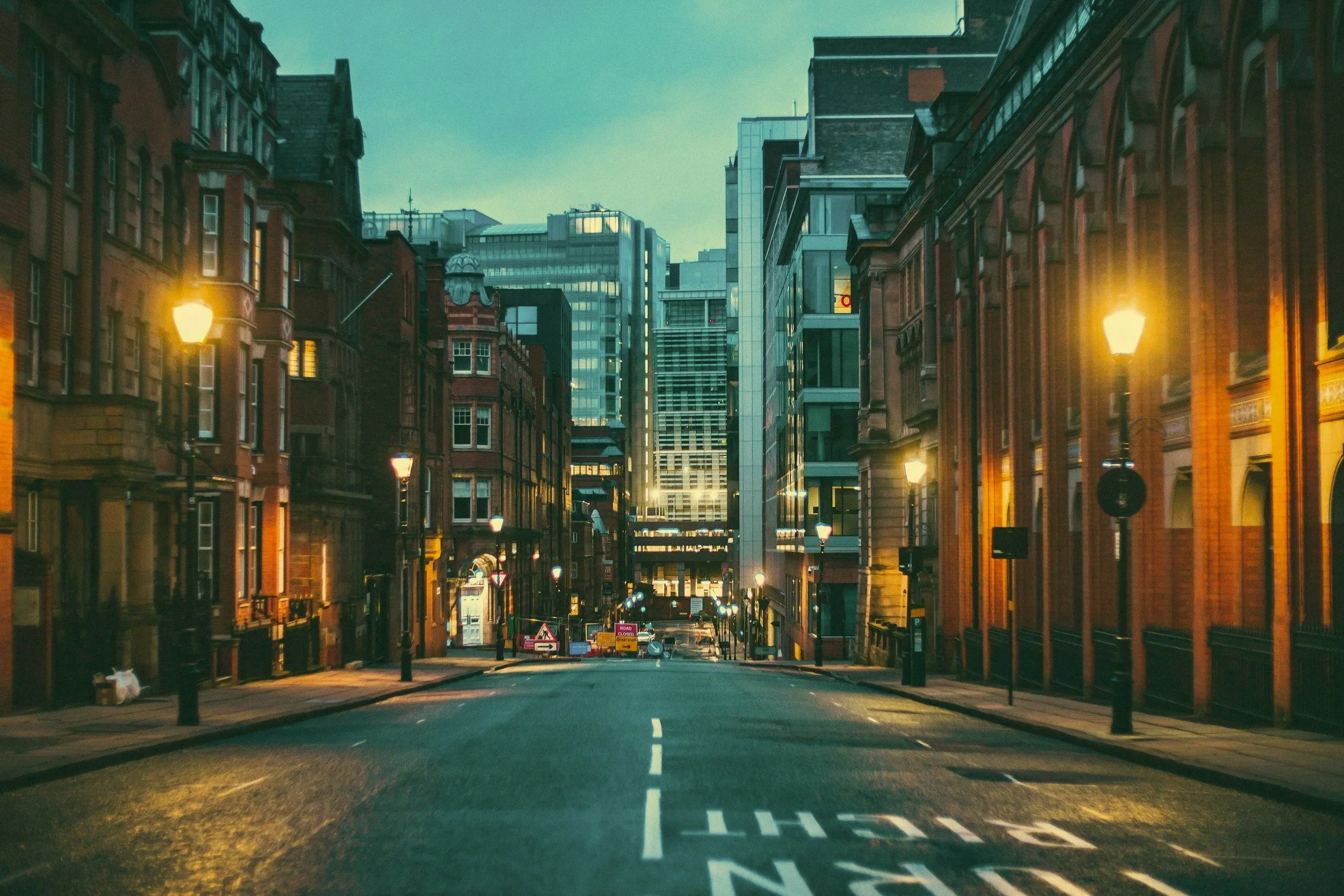 An empty city street at dusk with streetlights on flanking both sides, modern tall buildings in the background, and the road marked with a lane dividing line and a painted message.