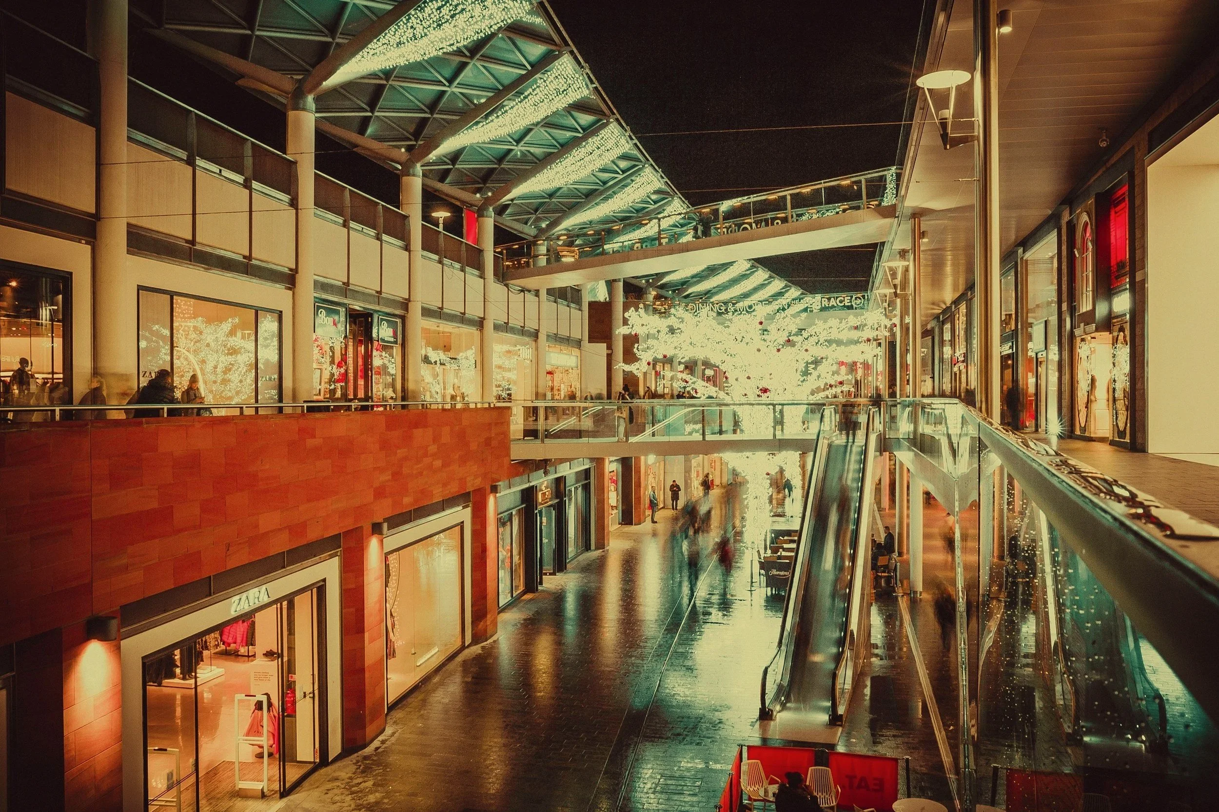 Interior of a shopping mall at night with lit Christmas decorations, escalators, and people walking.