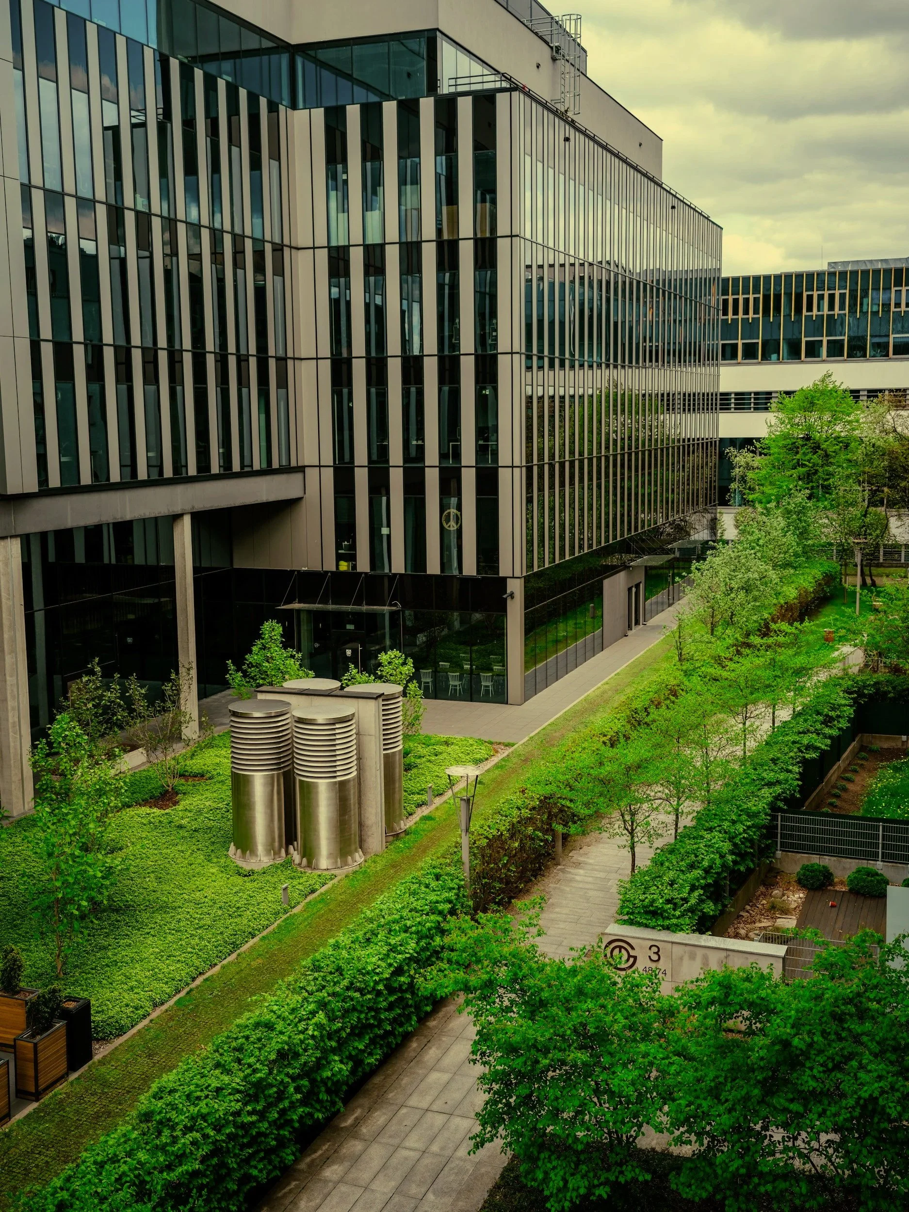 Modern office building with large glass windows, surrounded by green trees and landscaped outdoor area, including pathways and metallic ventilation structures.