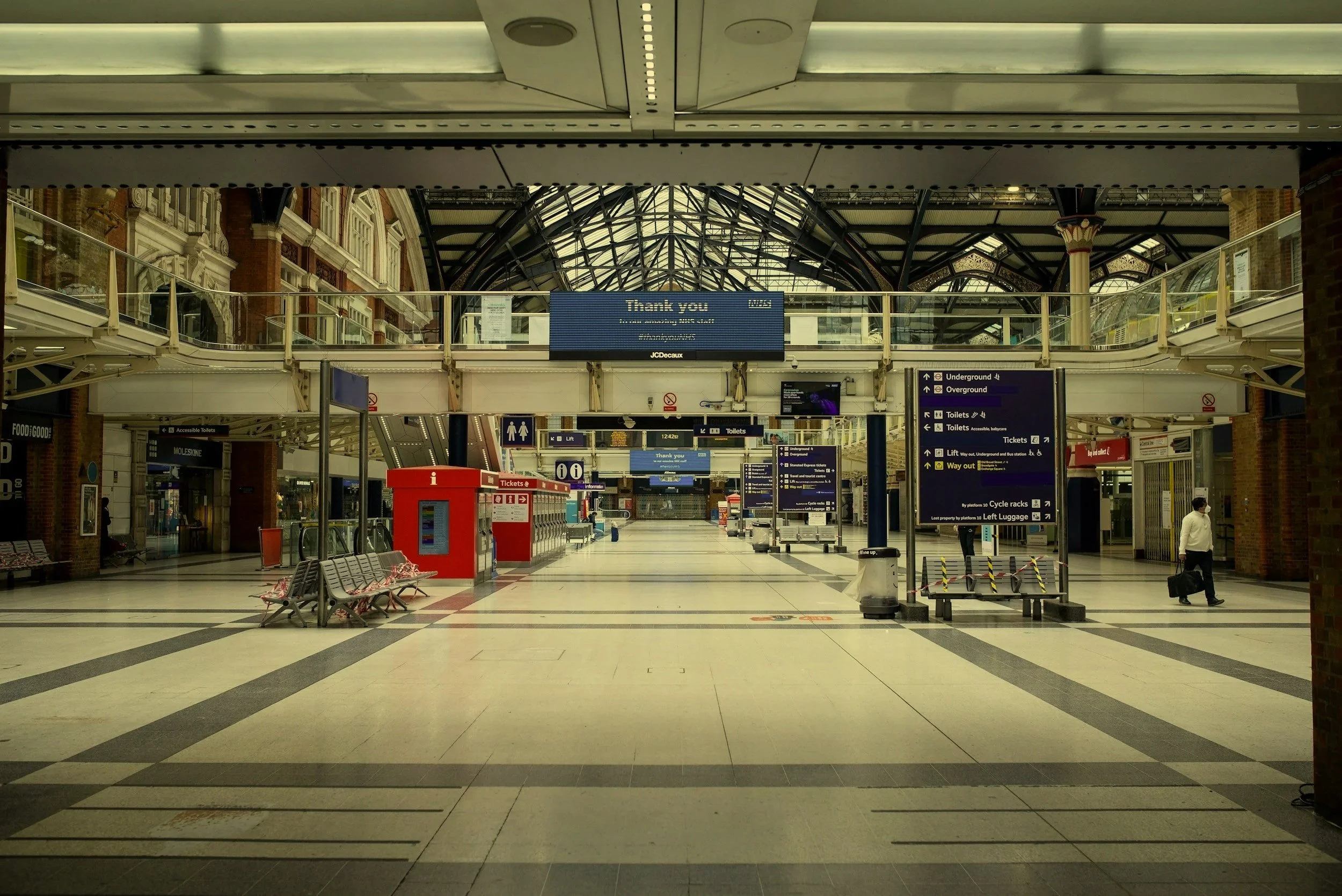 Empty train station interior with overhead signs, ticket machines, benches, and a few people walking with luggage.