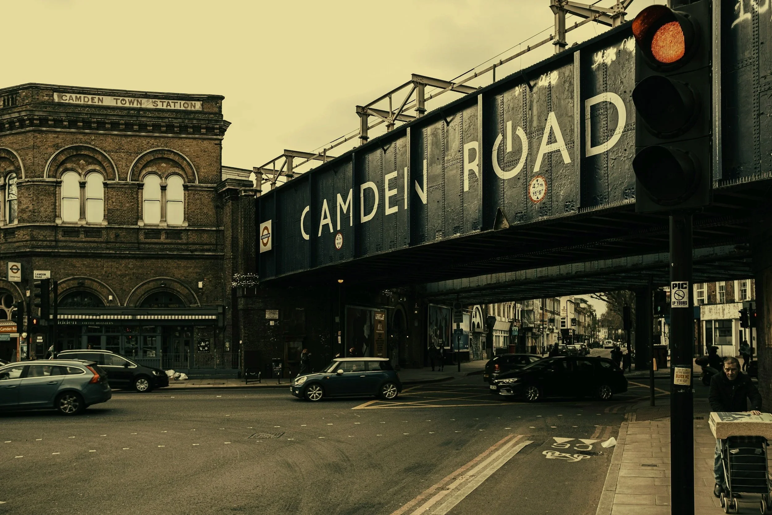 Street scene in London with cars, people, and a bridge with a sign labeled 'Camden Road' over a busy intersection, historic brick building in the background, and traffic lights.