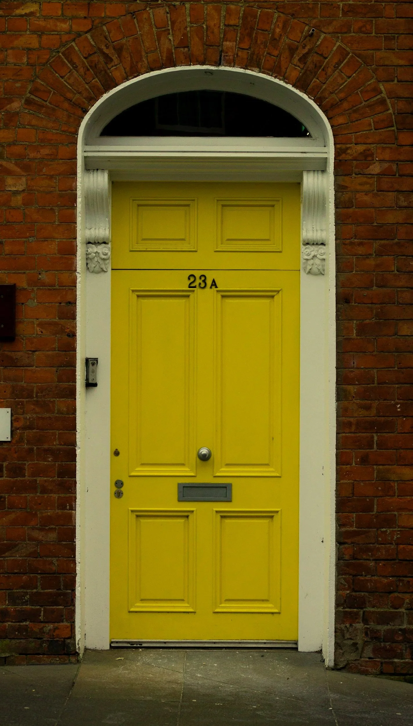 Bright yellow front door with decorative paneling, black house number '23A', a mail slot, a door handle, and a round doorknob, set in a red brick wall with a white frame and arched window above.