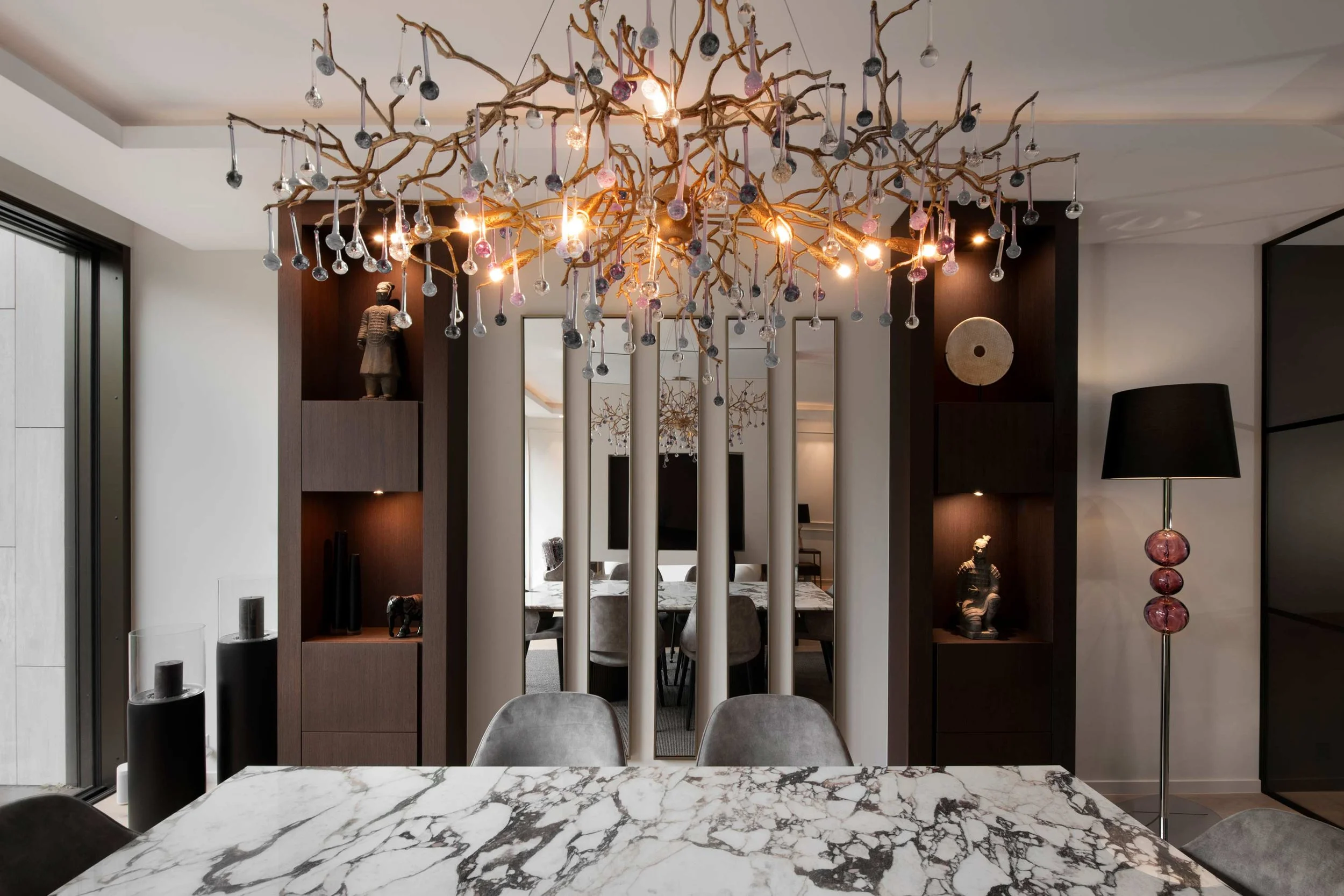Modern dining room with a marble table, decorative branch chandelier with hanging glass ornaments, mirrored wall panels, dark niche shelving with art objects, and a tall floor lamp with a black shade and pink glass base.