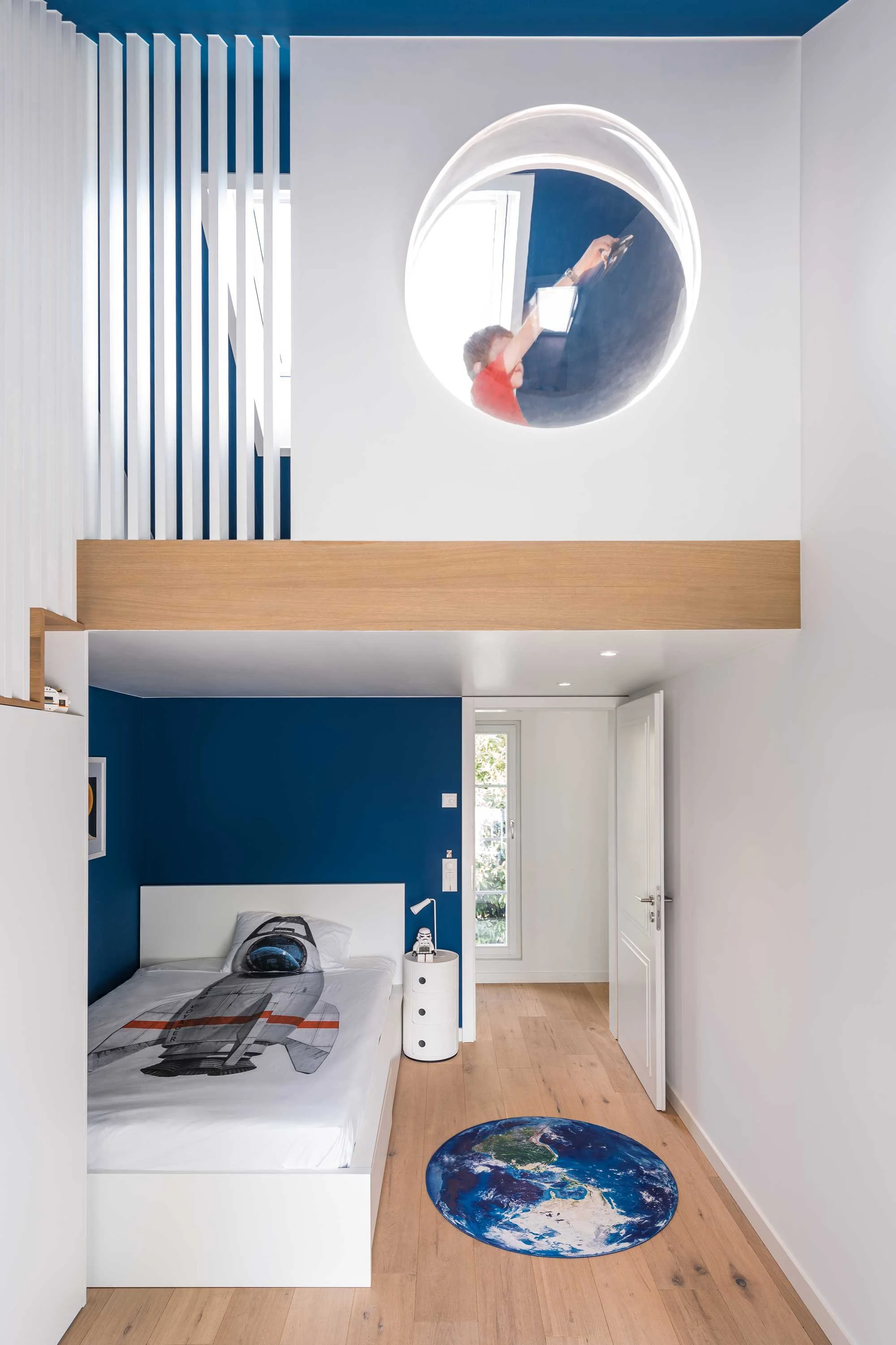 Children's bedroom with a blue accent wall, white bed with space-themed bedding, a round Earth-shaped rug, and a loft area with a large circular mirror reflecting a child.