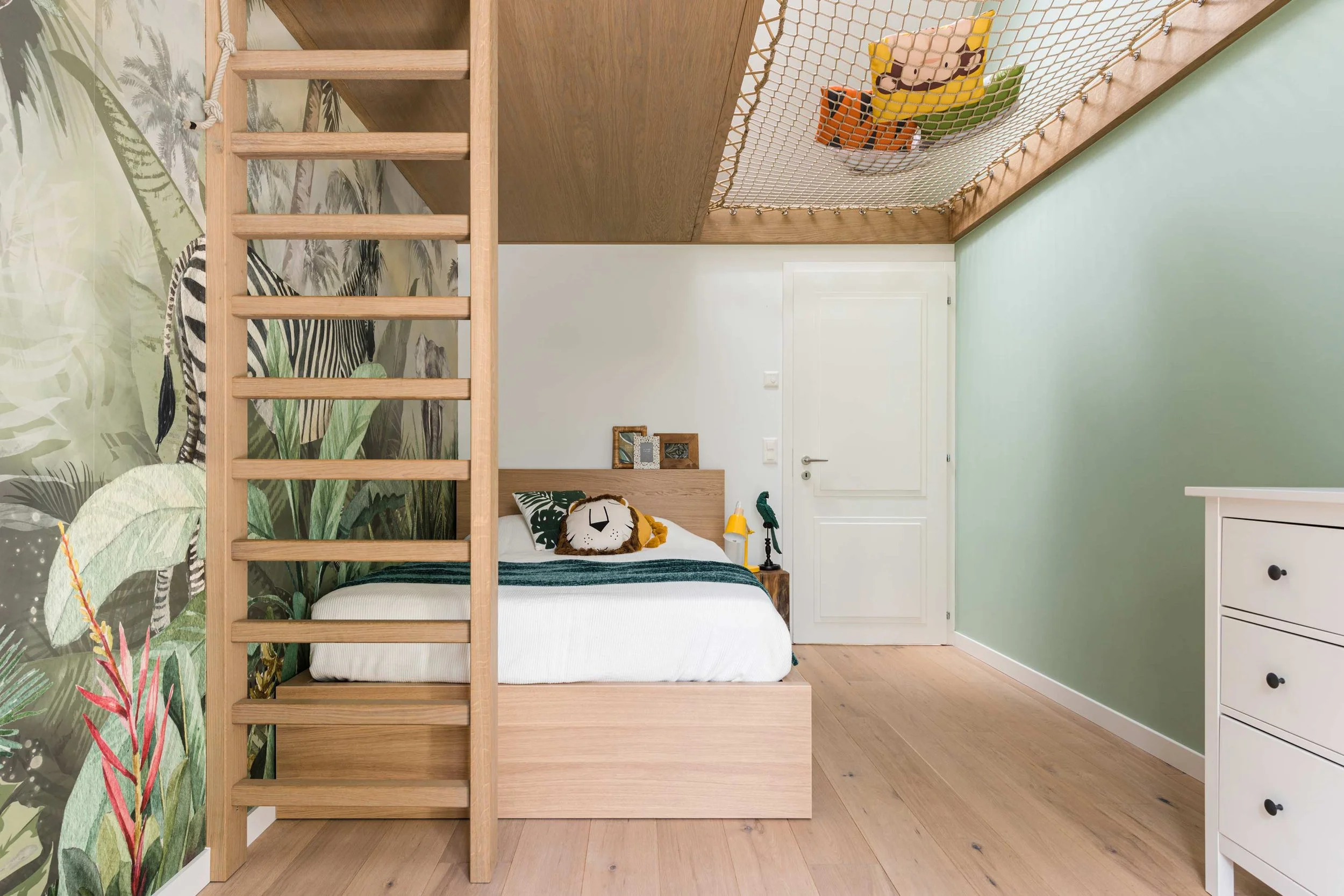 A bedroom with a jungle-themed wall mural, a wooden bed with a decorative lion pillow, a small white dresser, and a hanging netted section with toys above the bed.