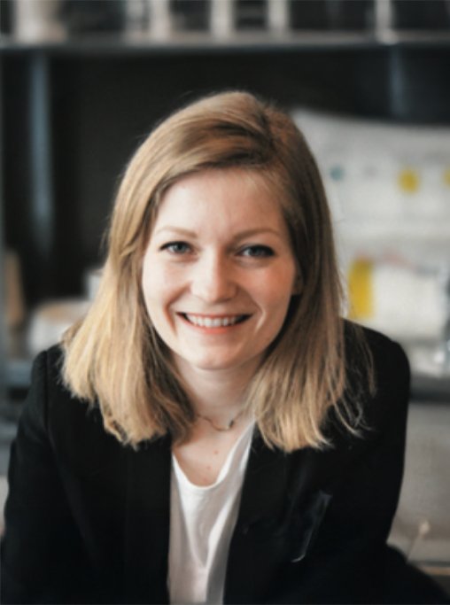 A young woman with blonde hair smiling at the camera, wearing a black blazer and white shirt, in an office or workspace setting.