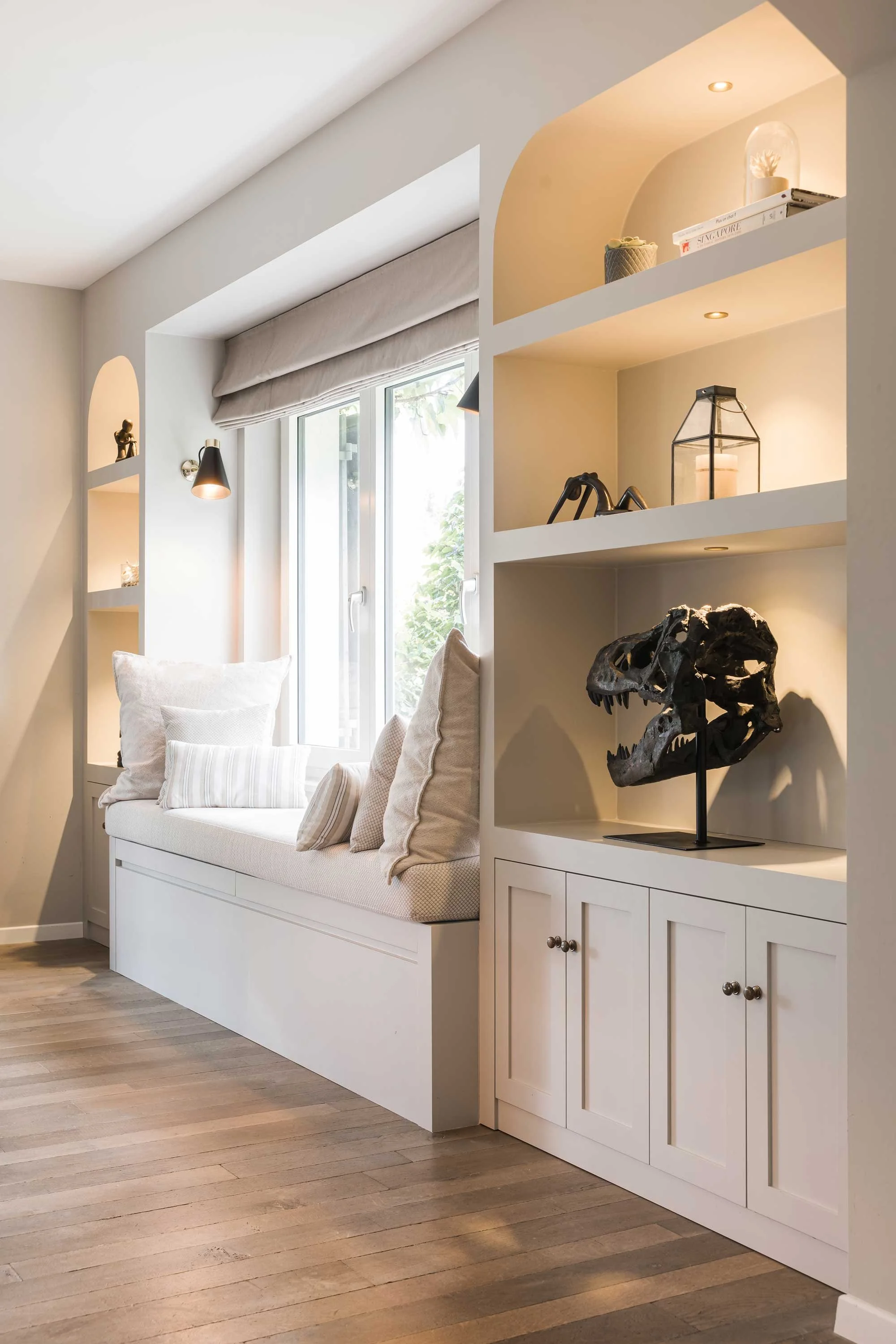 A cozy window nook with a cushioned bench and decorative pillows, situated next to built-in white shelves with books and decor, in a softly lit room with wooden flooring.