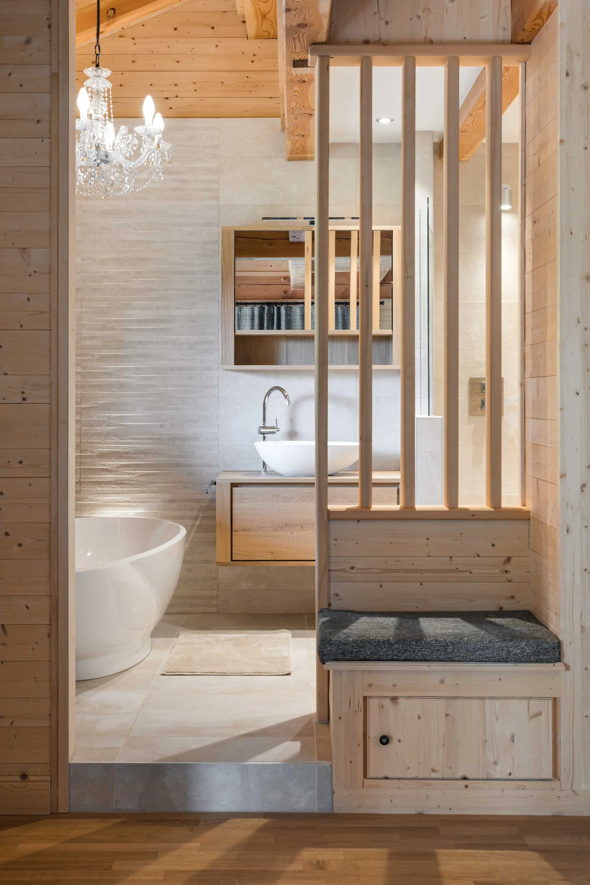 Interior view of a bathroom with wooden walls and ceiling, featuring a white freestanding bathtub, a floating wooden vanity with a vessel sink, a mirror, and a decorative chandelier hanging from the ceiling.
