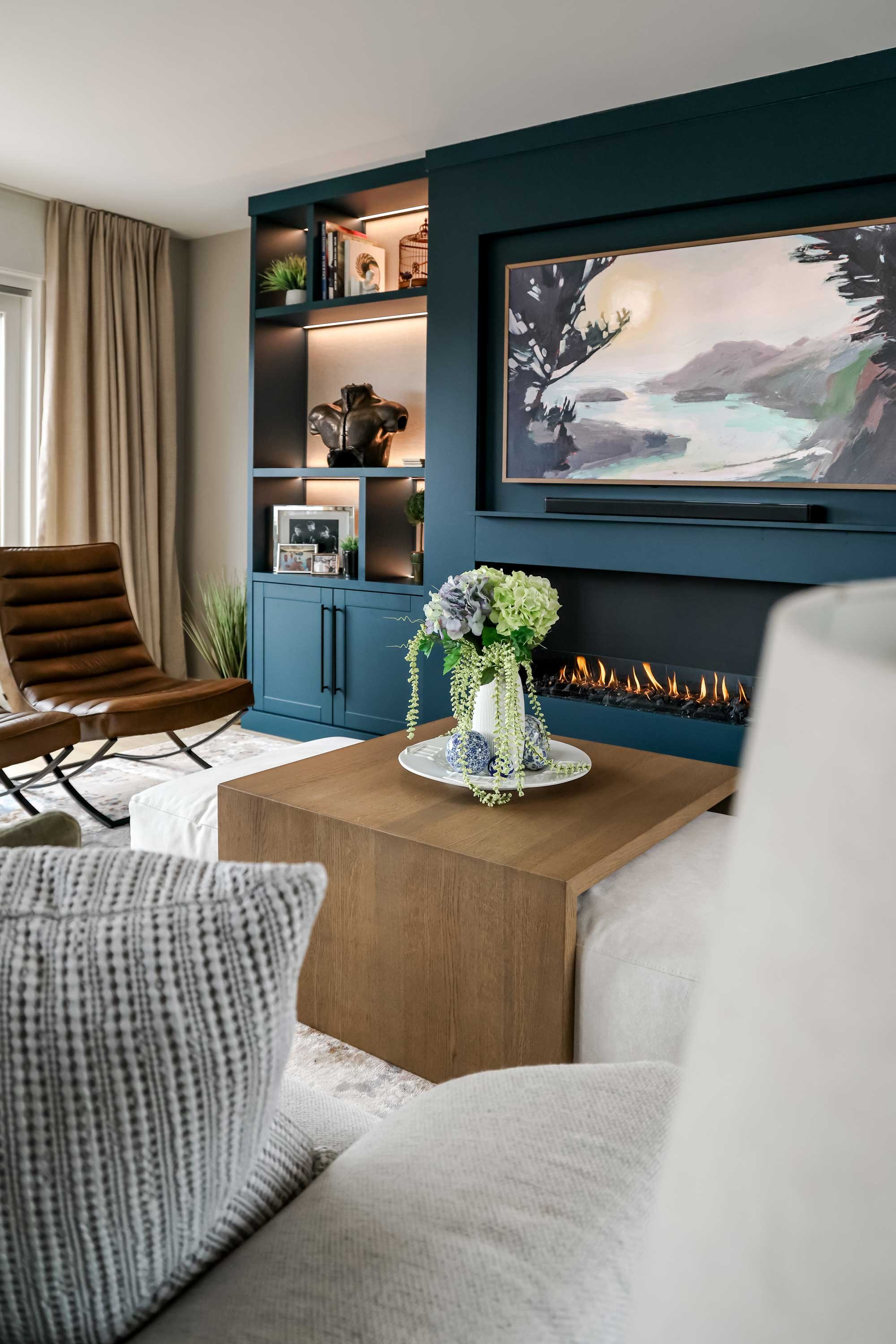Living room interior design with a beige sofa, a brown leather chair, a wooden coffee table with a flower arrangement, a wall-mounted electric fireplace, a large landscape painting, and a built-in blue cabinet with shelves and closed cabinets.