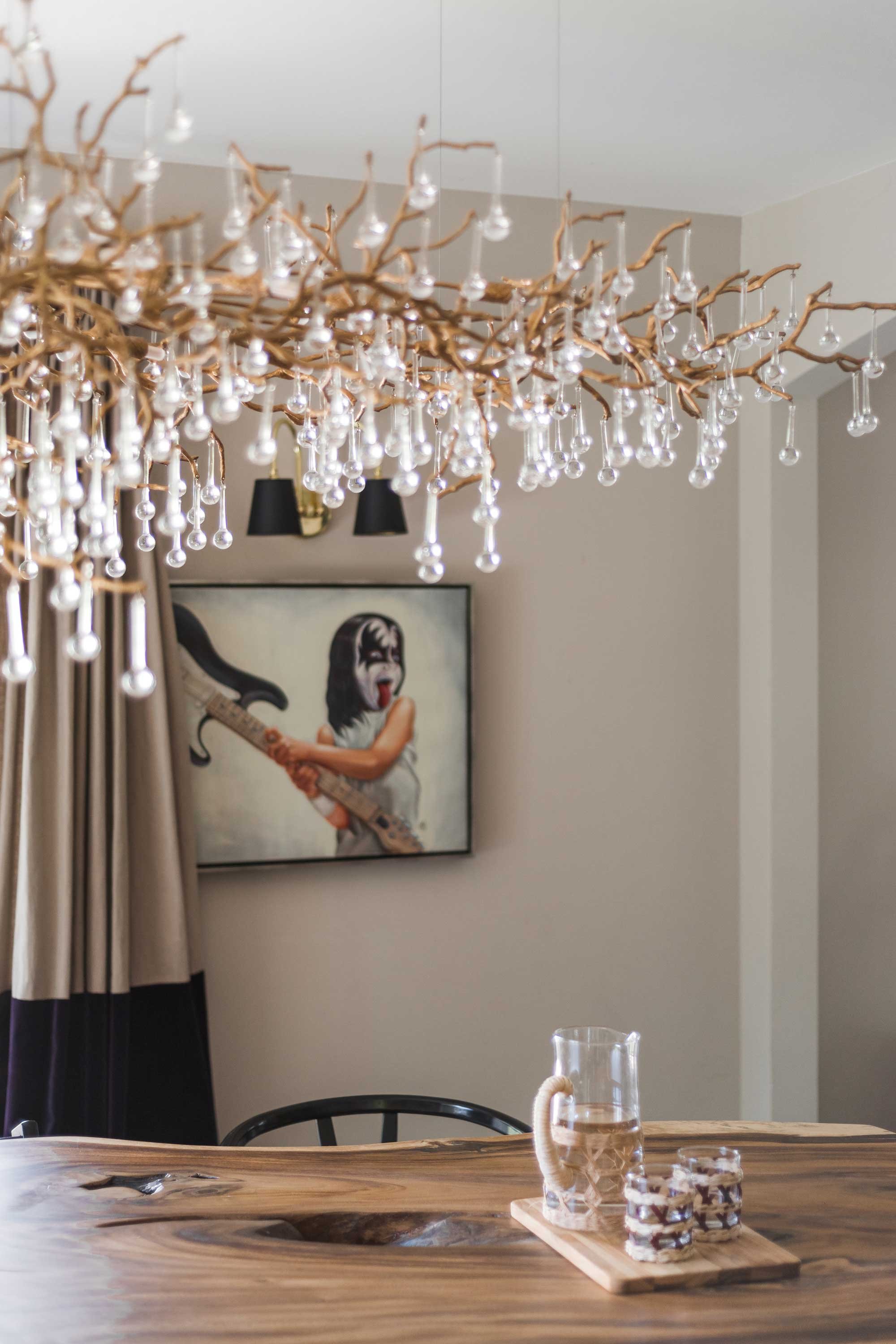 Interior dining room with a wooden table, glass pitcher of water, and two glasses on a wooden tray. A chandelier with glass droplets hangs above. A framed artwork of a girl holding an electric guitar with face paint, with black and beige curtains in the background.