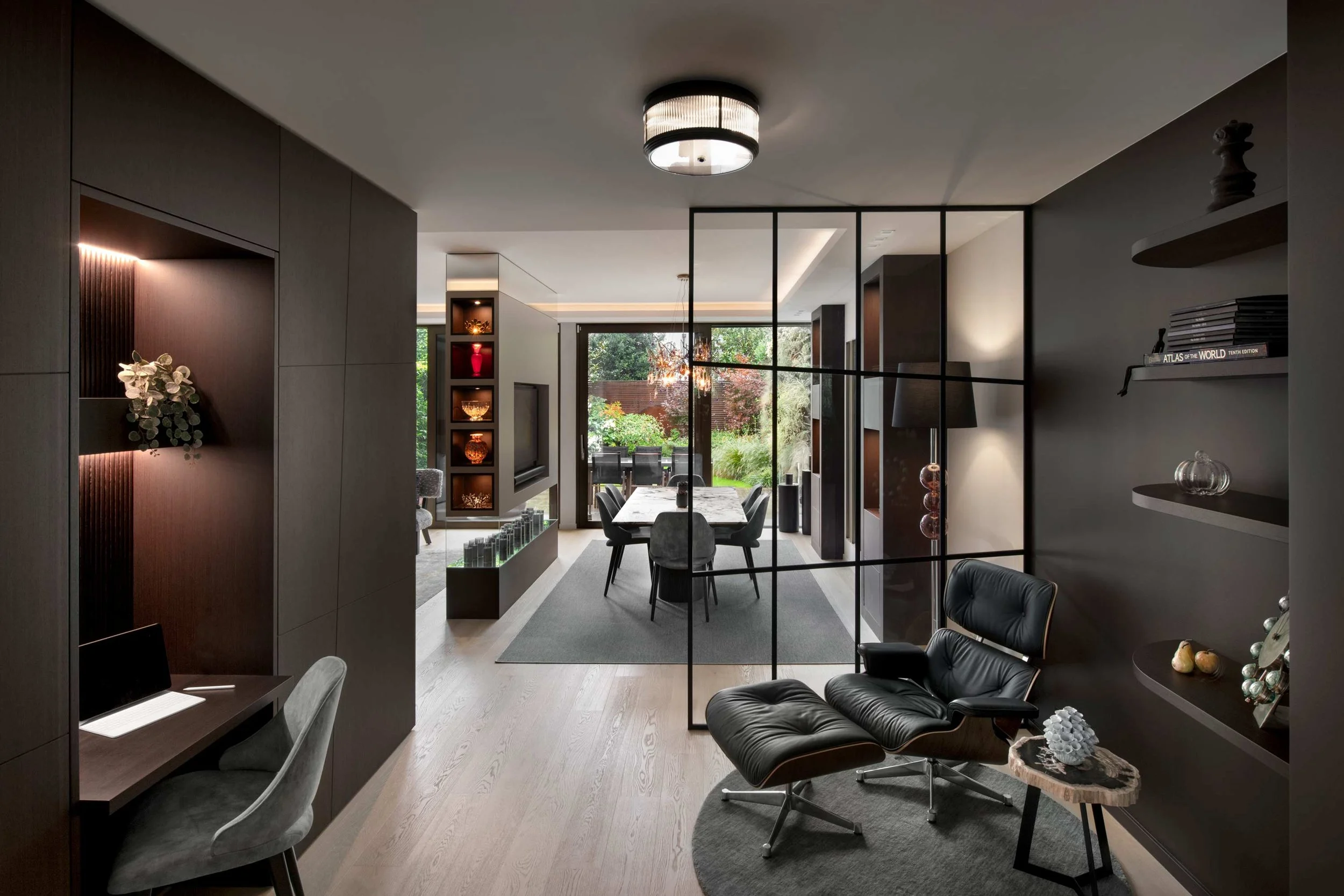Modern living room with a dining area, black leather lounge chair, shelves with books and decorative items, large windows with outdoor greenery, and contemporary lighting fixtures.