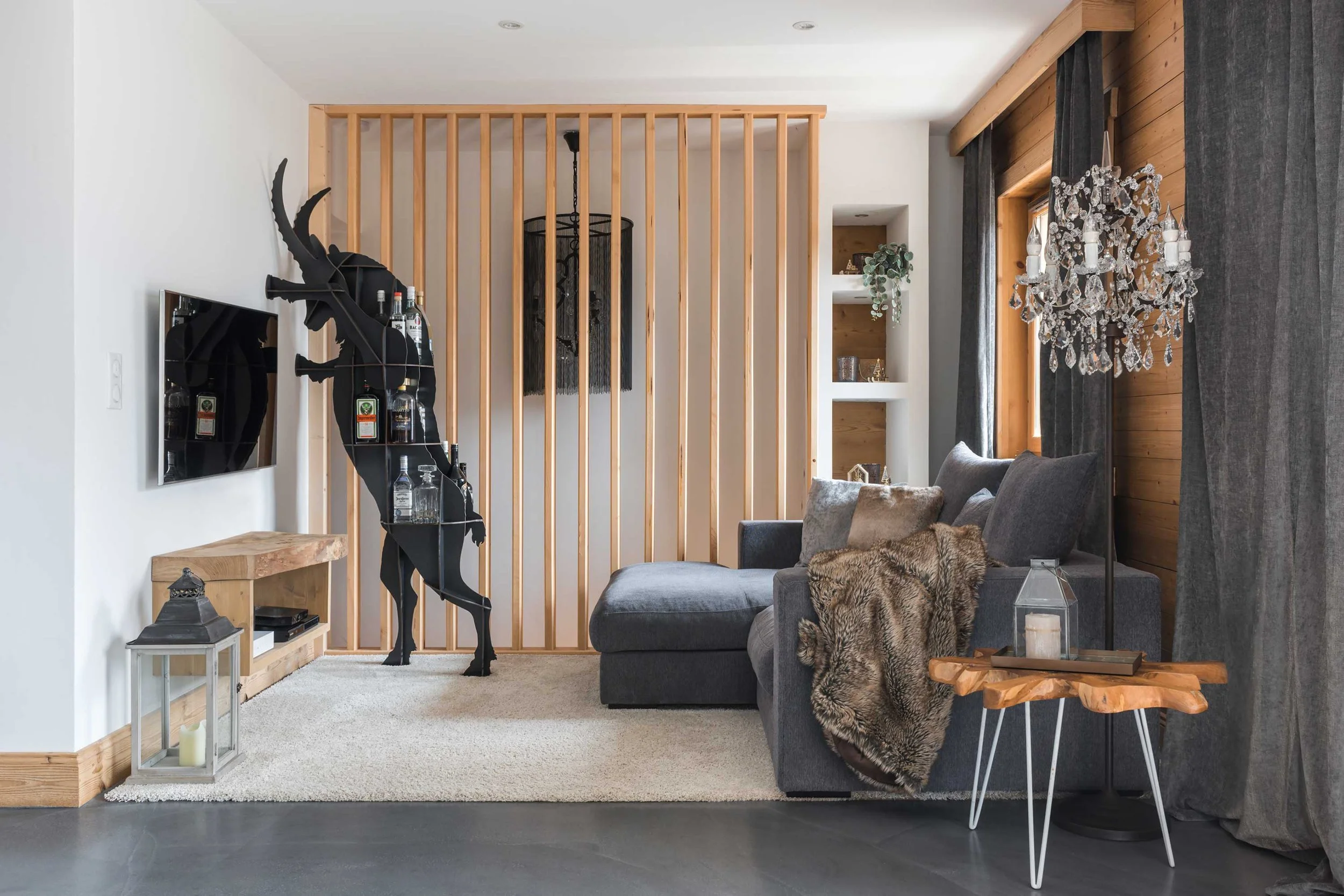Living room with gray sectional sofa, fur throw blanket, crystal chandelier, wooden side table, built-in shelves with decor, dark curtains, and a black bar sculpture of a person holding bottles.