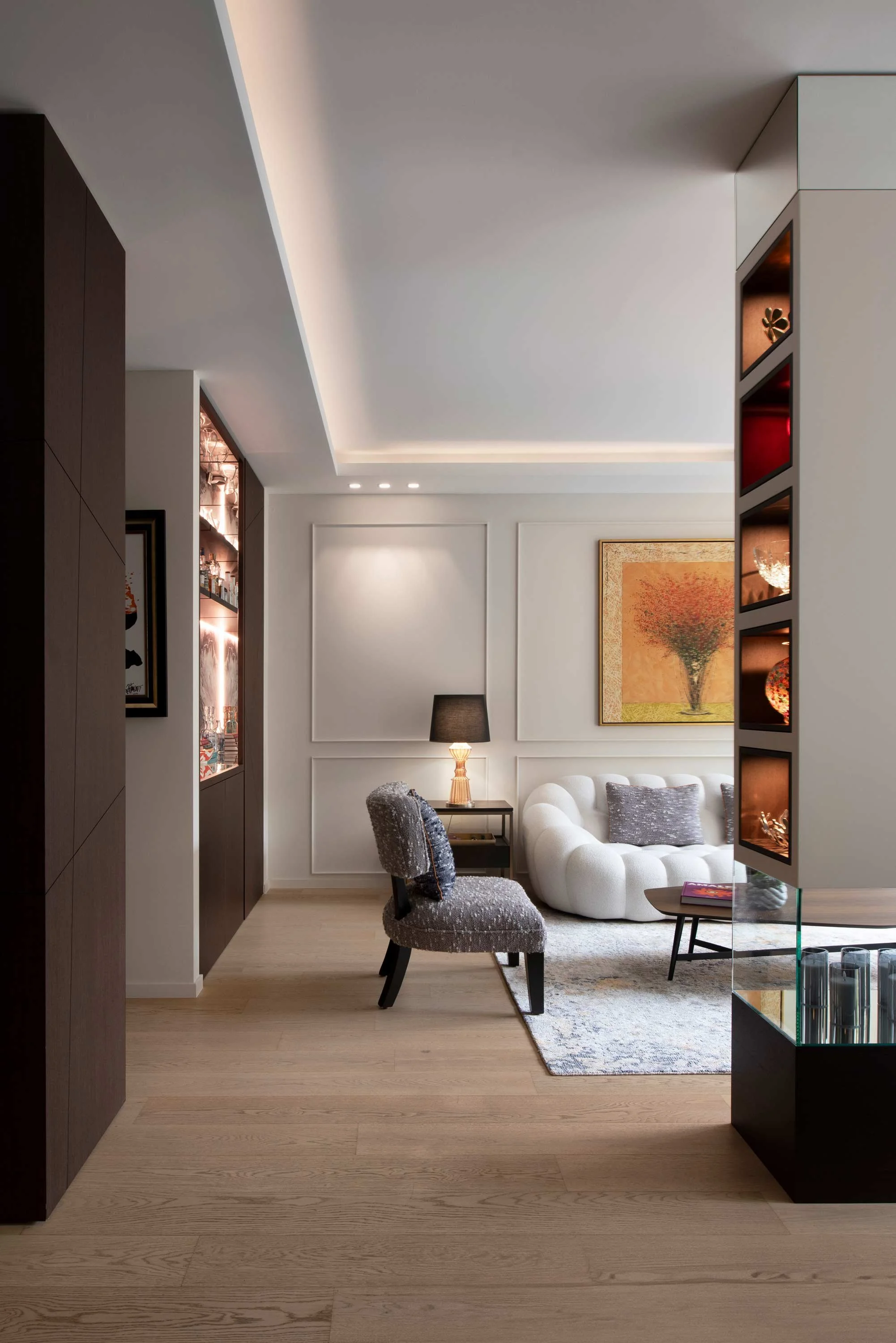 Modern living room with white sofa, gray armchair, wooden side table with lamp, wall art, and decorative display shelves in the background.