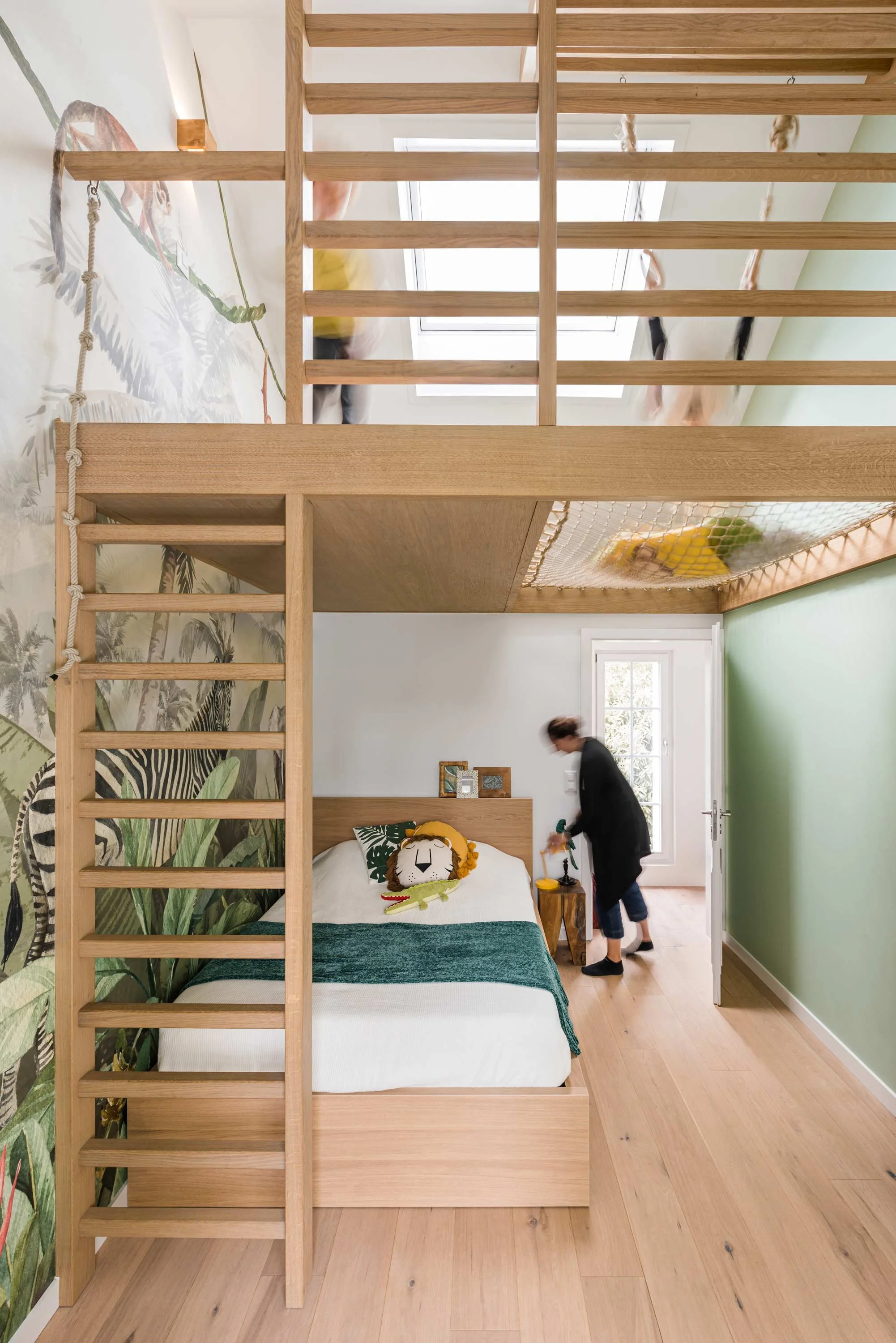 A bedroom with a bed featuring a lion plush toy and green blanket, wooden furniture, and a wall mural of jungle leaves and animals. An adult is standing near the door, and there's a loft with netting and a slide leading up, with blurred movement of a child on the slide.