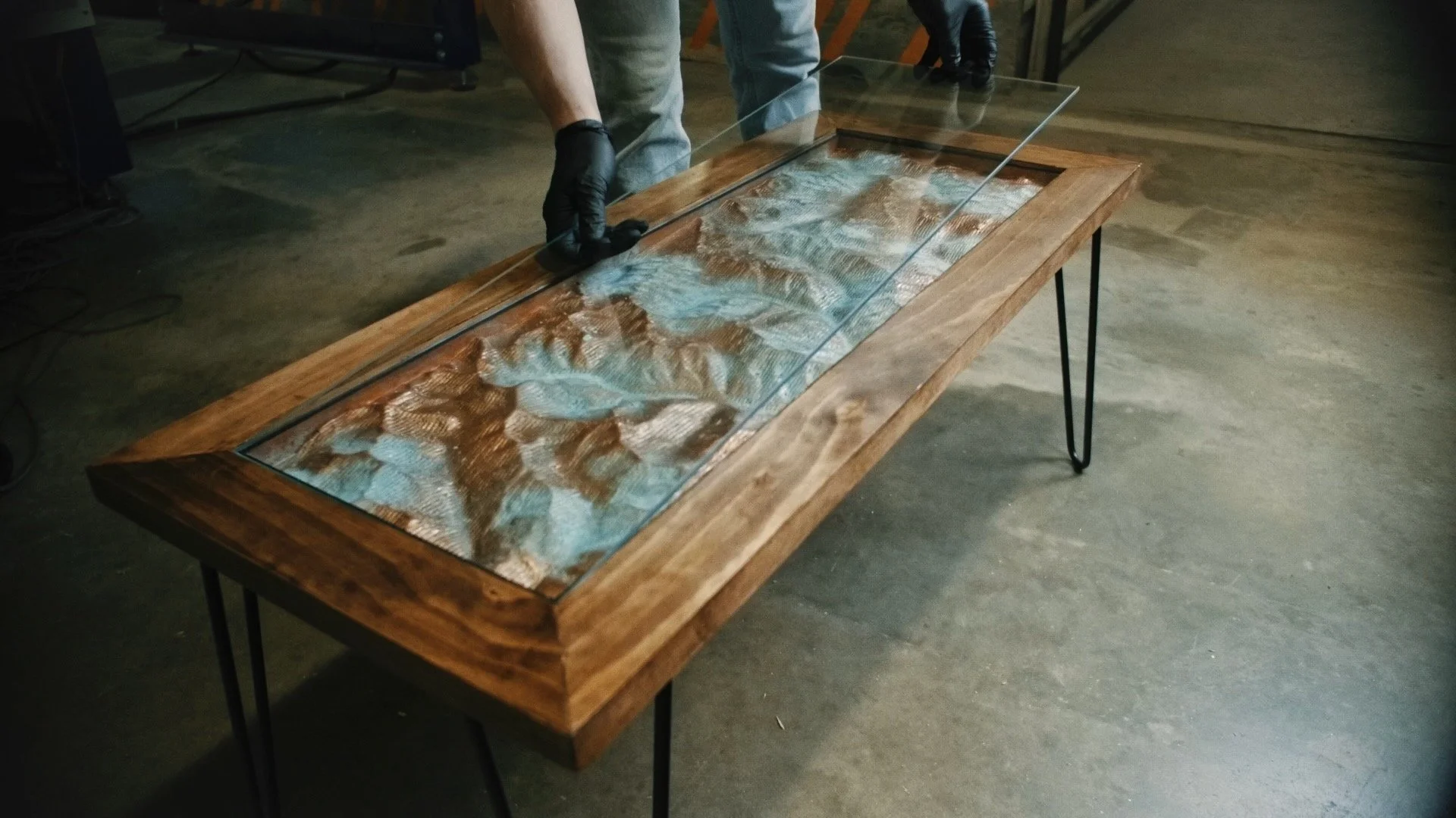 Person wearing black gloves is installing a glass top on a wooden table with a wooden frame and metal legs. The table has a decorative natural wood edge and an artistic resin surface underneath the glass.