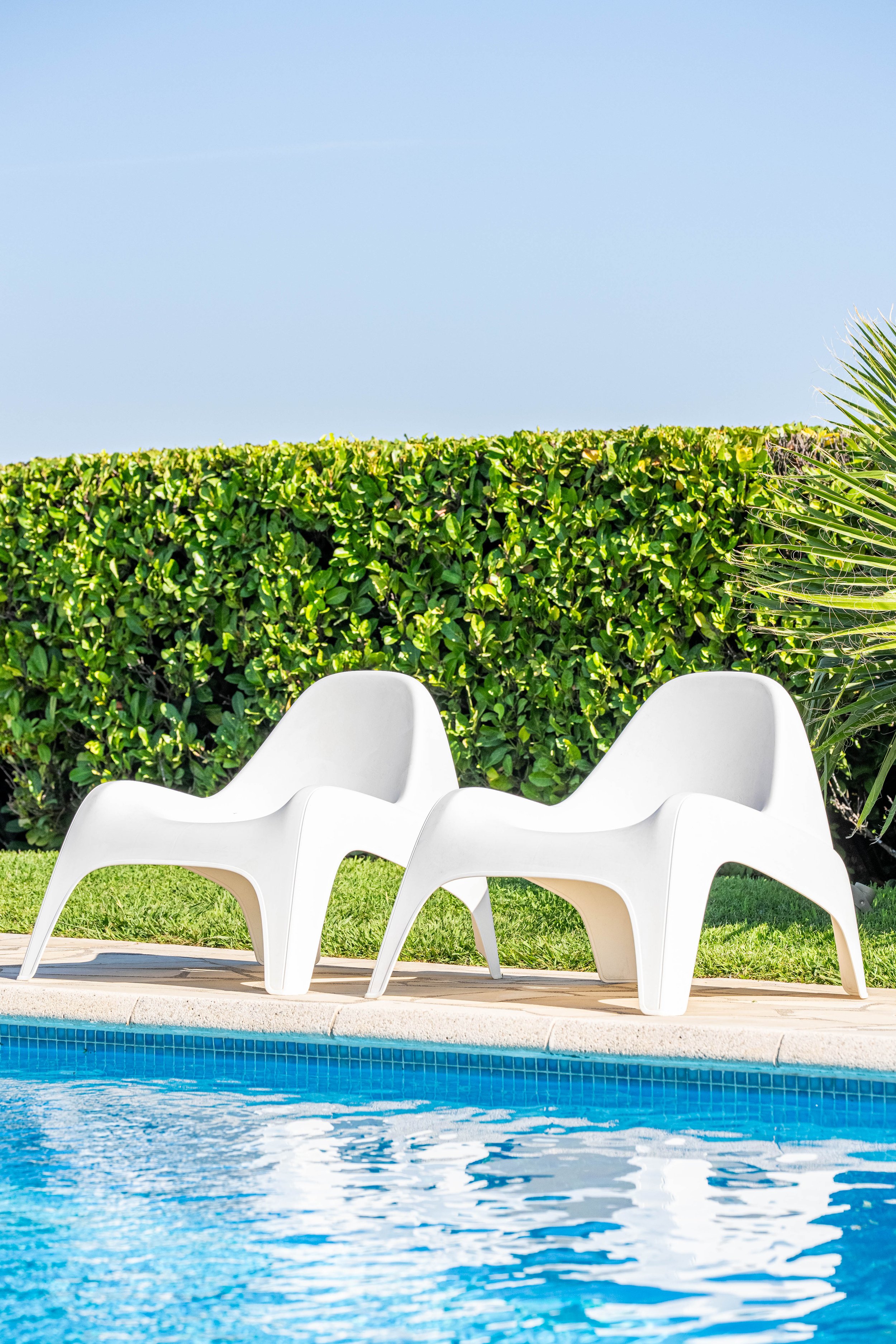 Chaises de plage blanches au bord de la piscine, avec un buisson verdoyant en arrière-plan