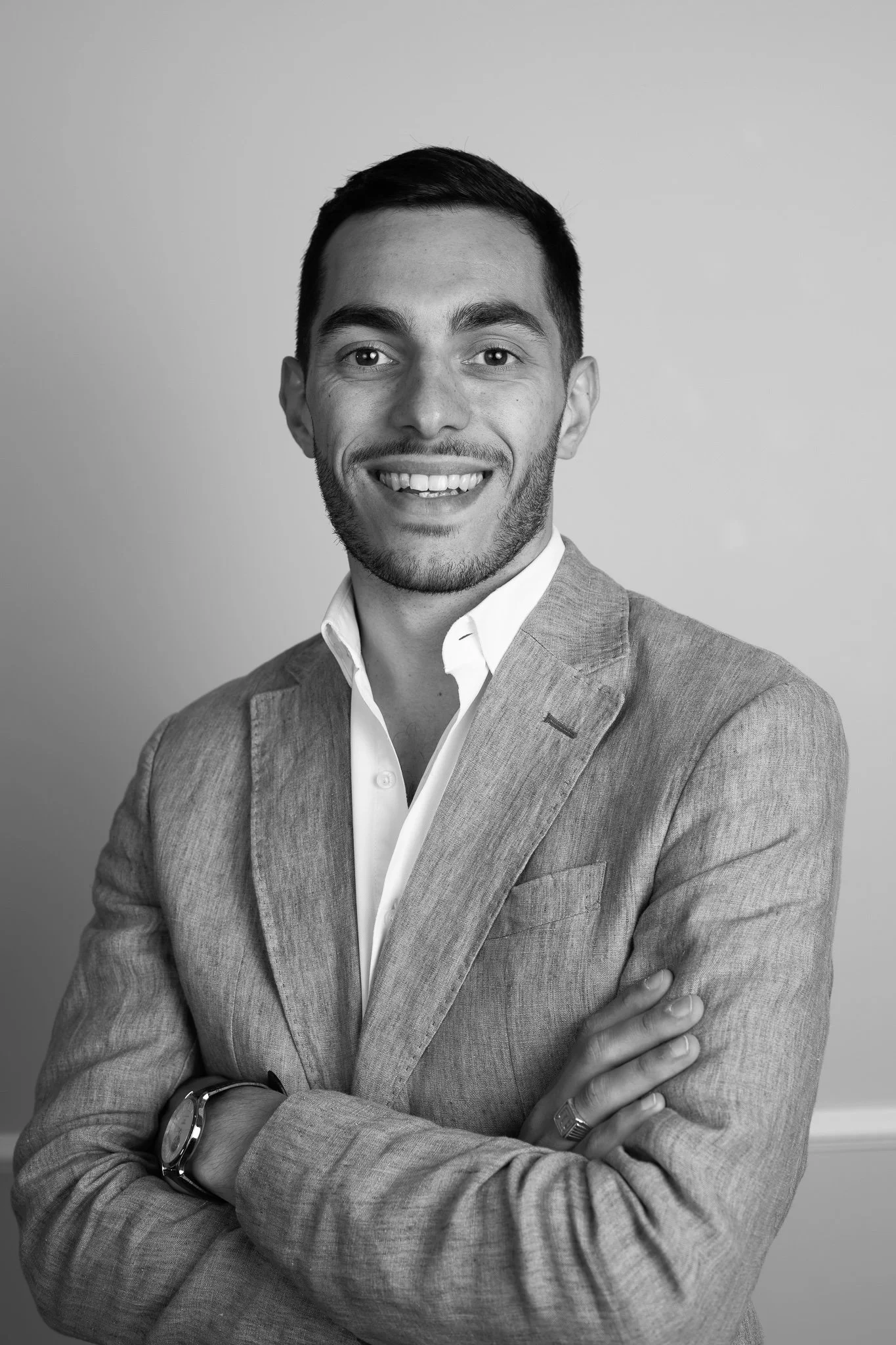 Portrait en noir et blanc d'un homme souriant, portant une veste en tissu et une chemise blanche, bras croisés, regardant vers l'appareil.