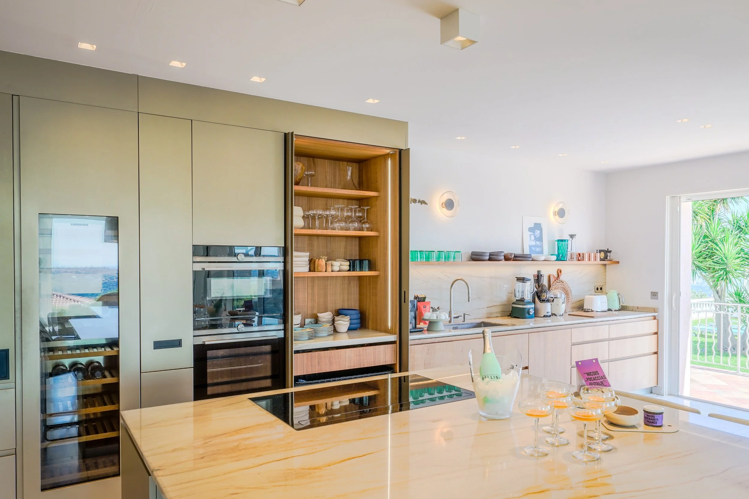 Cuisine moderne lumineuse avec plan de travail en marbre, étagères en bois, appareils électroménagers, bouteille de champagne dans un seau à glace, et balcon en arrière-plan avec vue sur la nature
