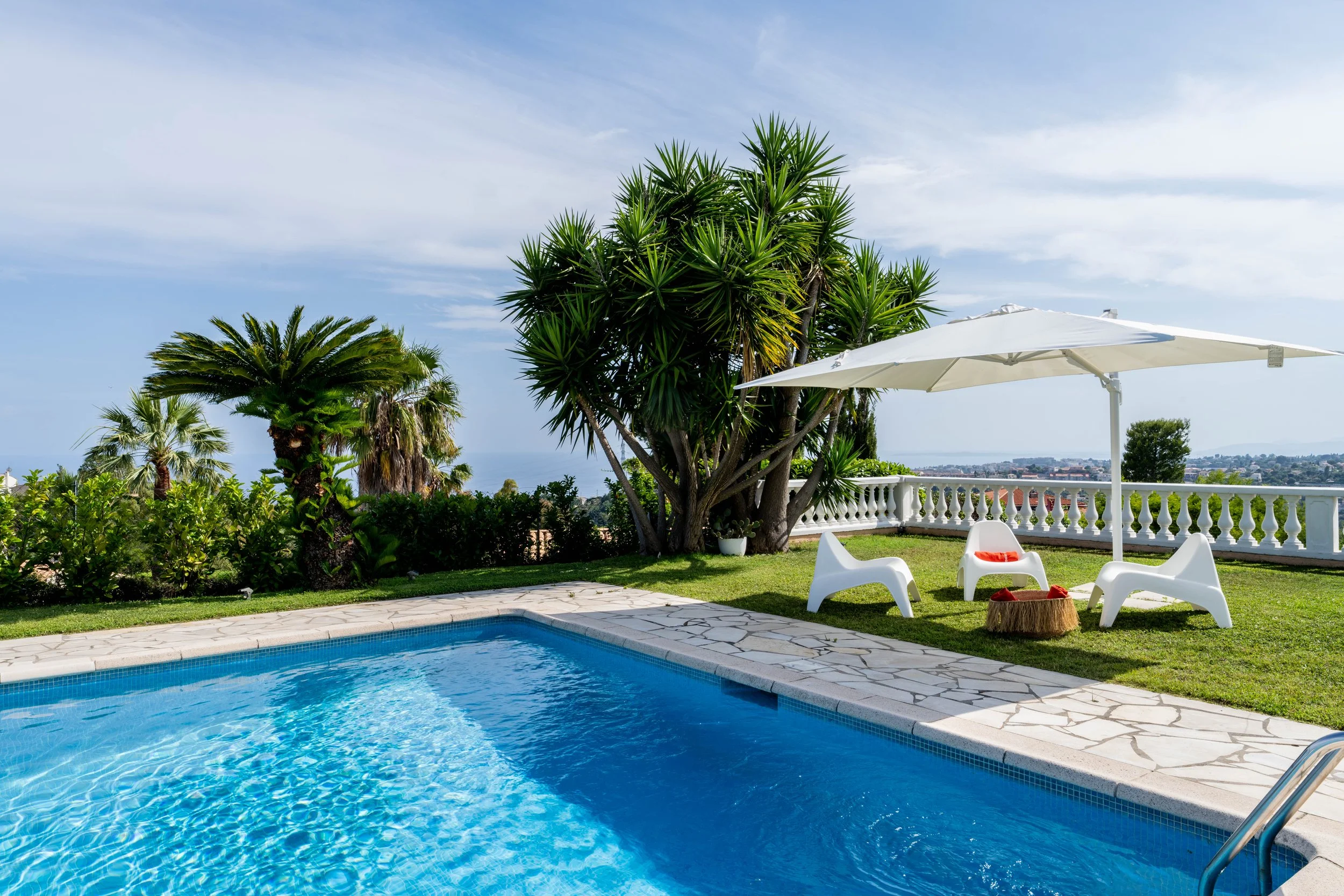 Piscine extérieure avec mobilier de détente sous un parasol, entourée de végétation tropicale et offrant une vue sur la mer