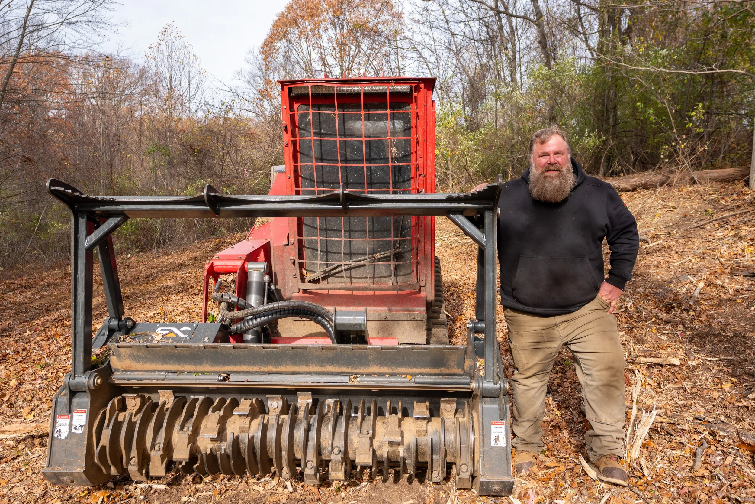 A man with a beard, black hoodie, and tan pants standing next to a red skid steer loader on a leaf-covered wooded hillside.