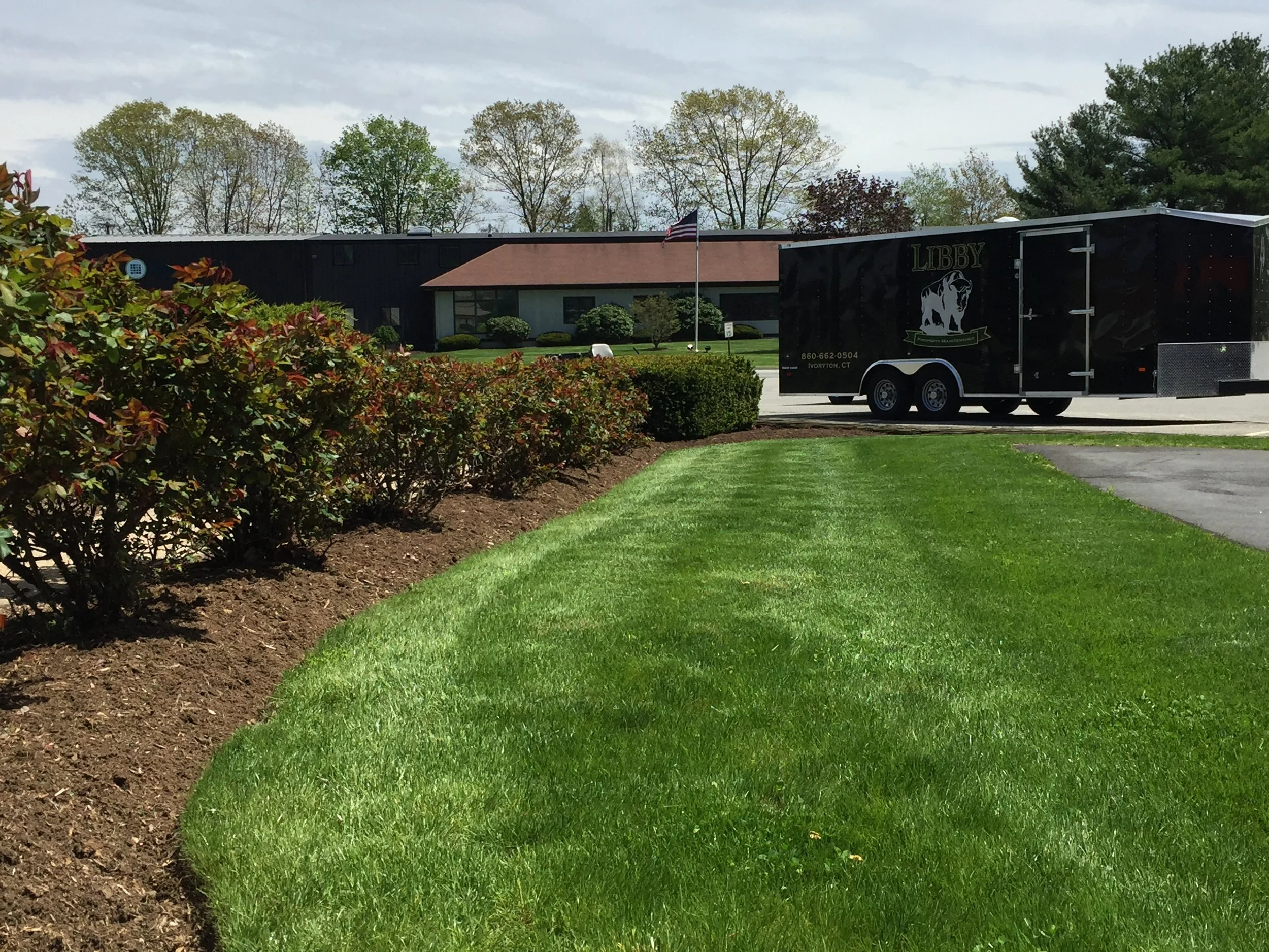 A well-maintained lawn and bushes in front of a building with a parking lot. A black trailer with the word "LIBBY" and a bulldog logo is parked on the asphalt.