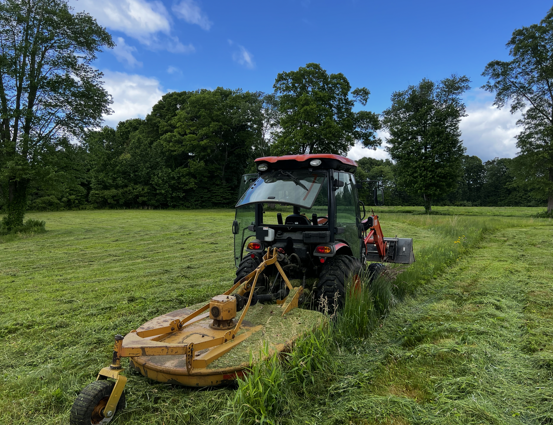 A tractor mowing grass in a scenic rural area with green trees and a blue sky with some clouds.