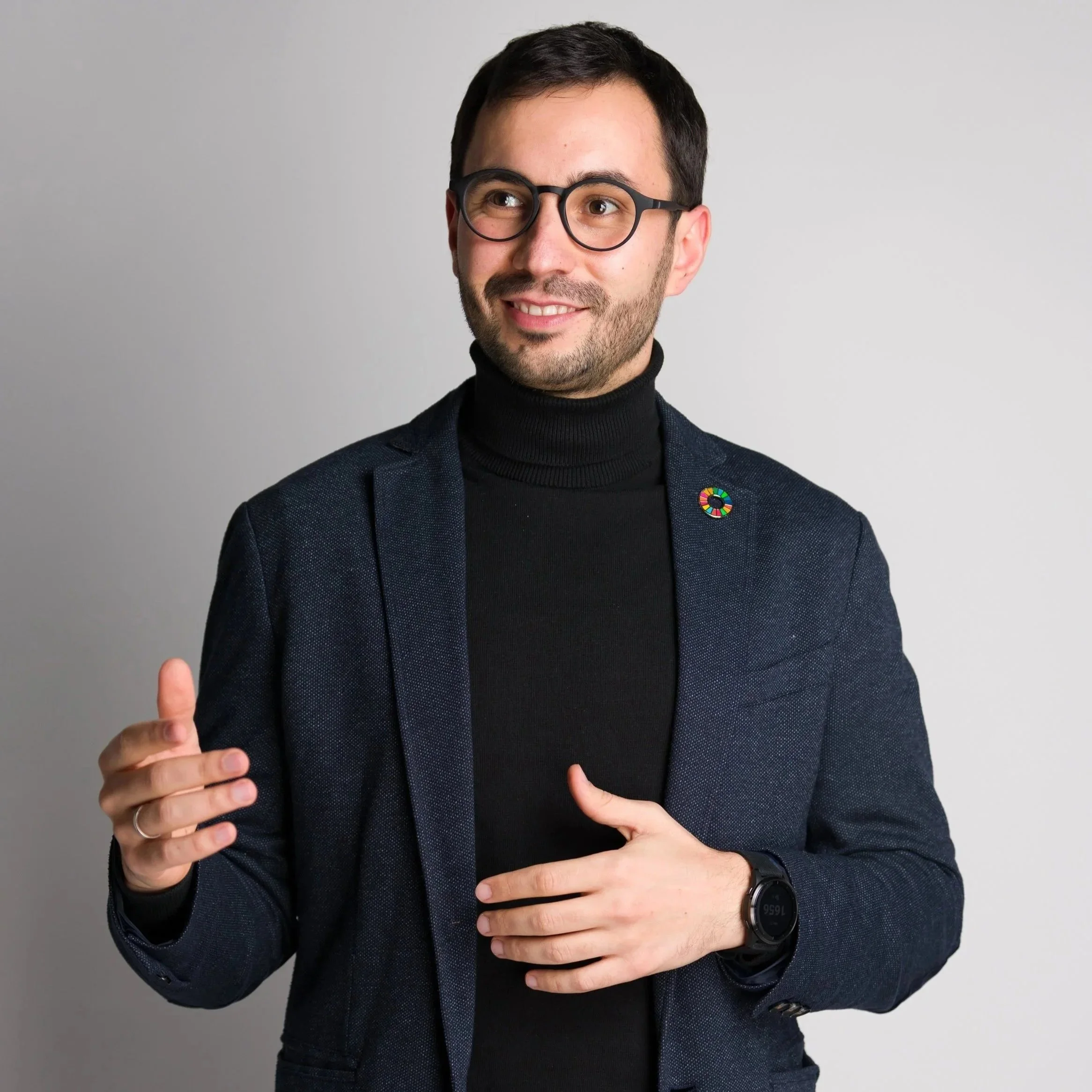 Tudor Tarlev - the founder of omcanvas. Wearing glasses, a black turtleneck, and a dark blazer with a United Nations Sustainable Development Goals pin, smiling and gesturing with his right hand in front of a plain light gray background.