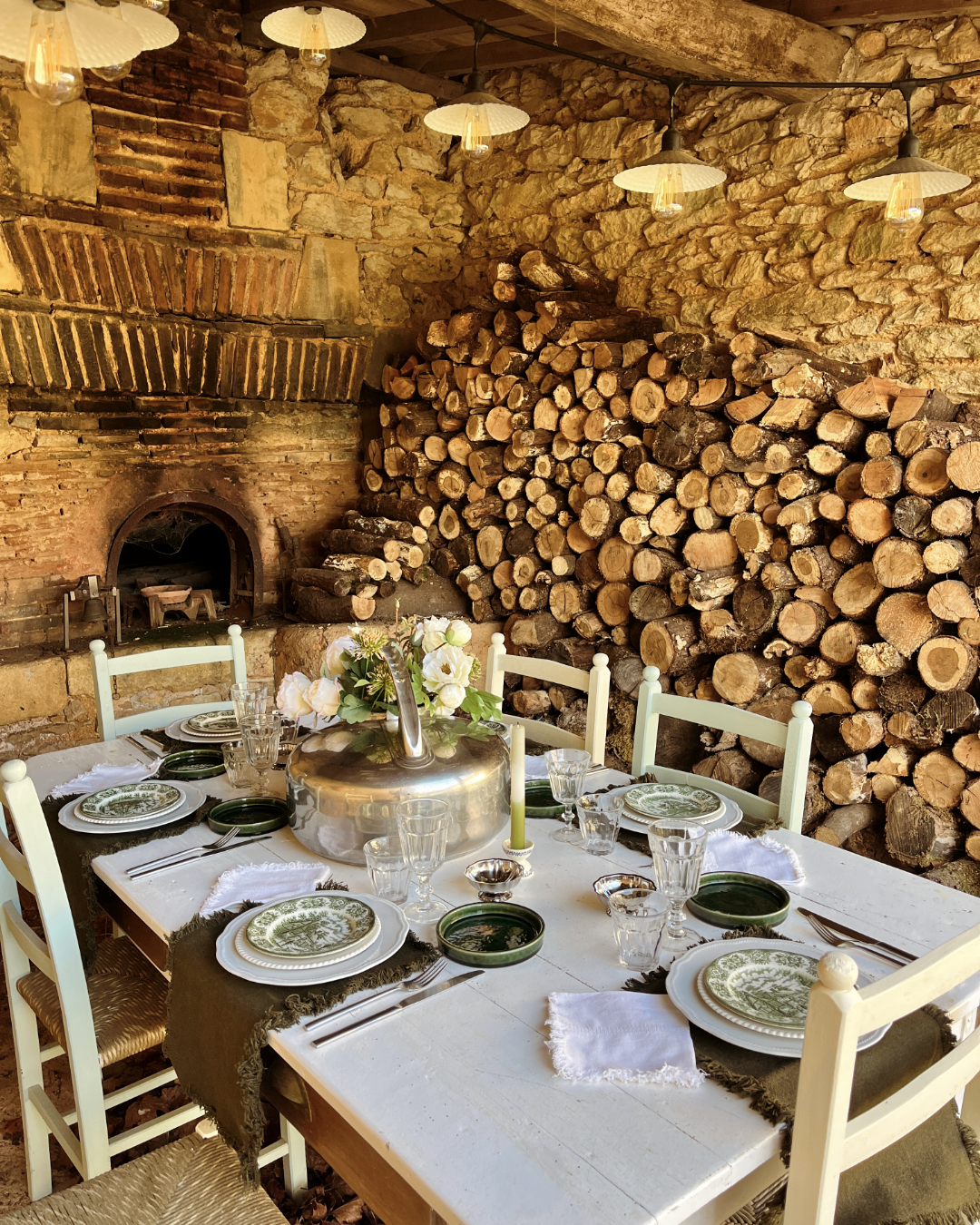Table de dîner dans une pièce rustique avec mur en pierre, cheminée en briques, et une pile de bois de chauffage. La table est décorée avec un vase de fleurs, des assiettes en porcelaine, des verres, et des napperons, prête pour un repas convivial.