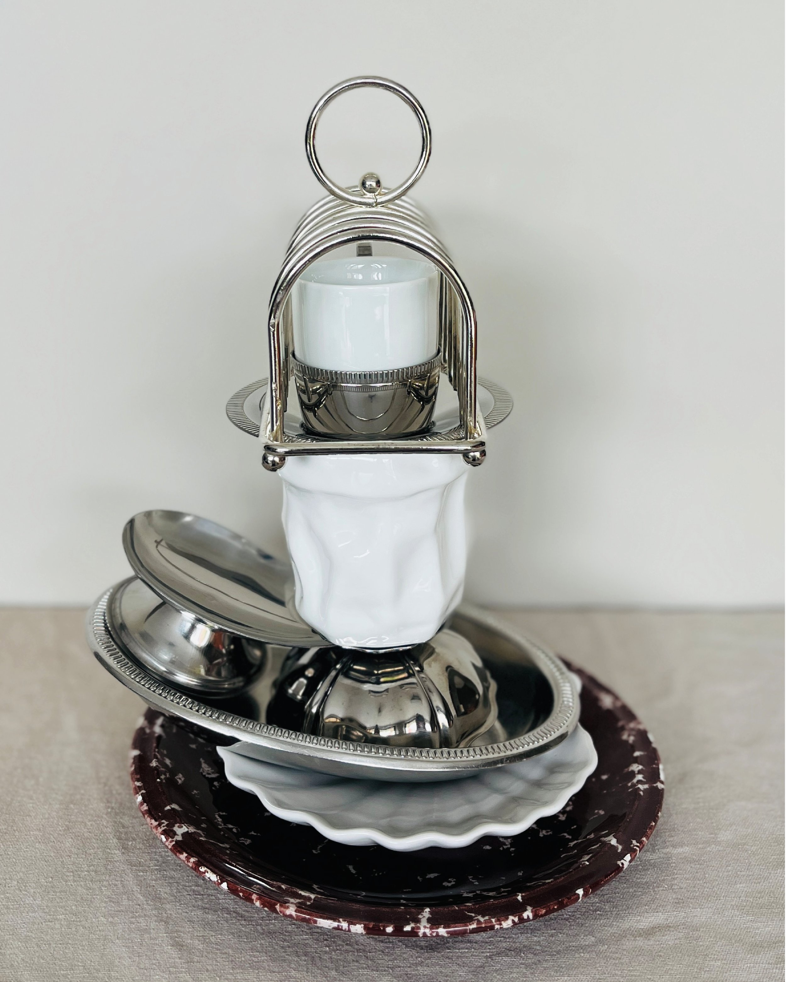 Une composition artistique d'objets de vaisselle en métal et céramique, formant une sculpture abstraite sur une assiette en céramique rouge avec une banderole blanche en arrière-plan.