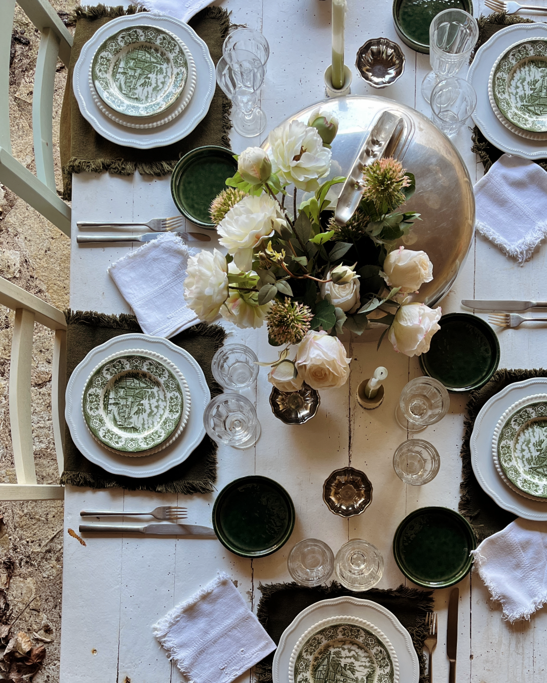 Table de dîner élégante avec vaisselle en porcelaine blanche et motifs verts, verres à vin et à eau, couverts en métal argenté, napperons en tissu blanc, bougies et un vase avec des fleurs blanches.