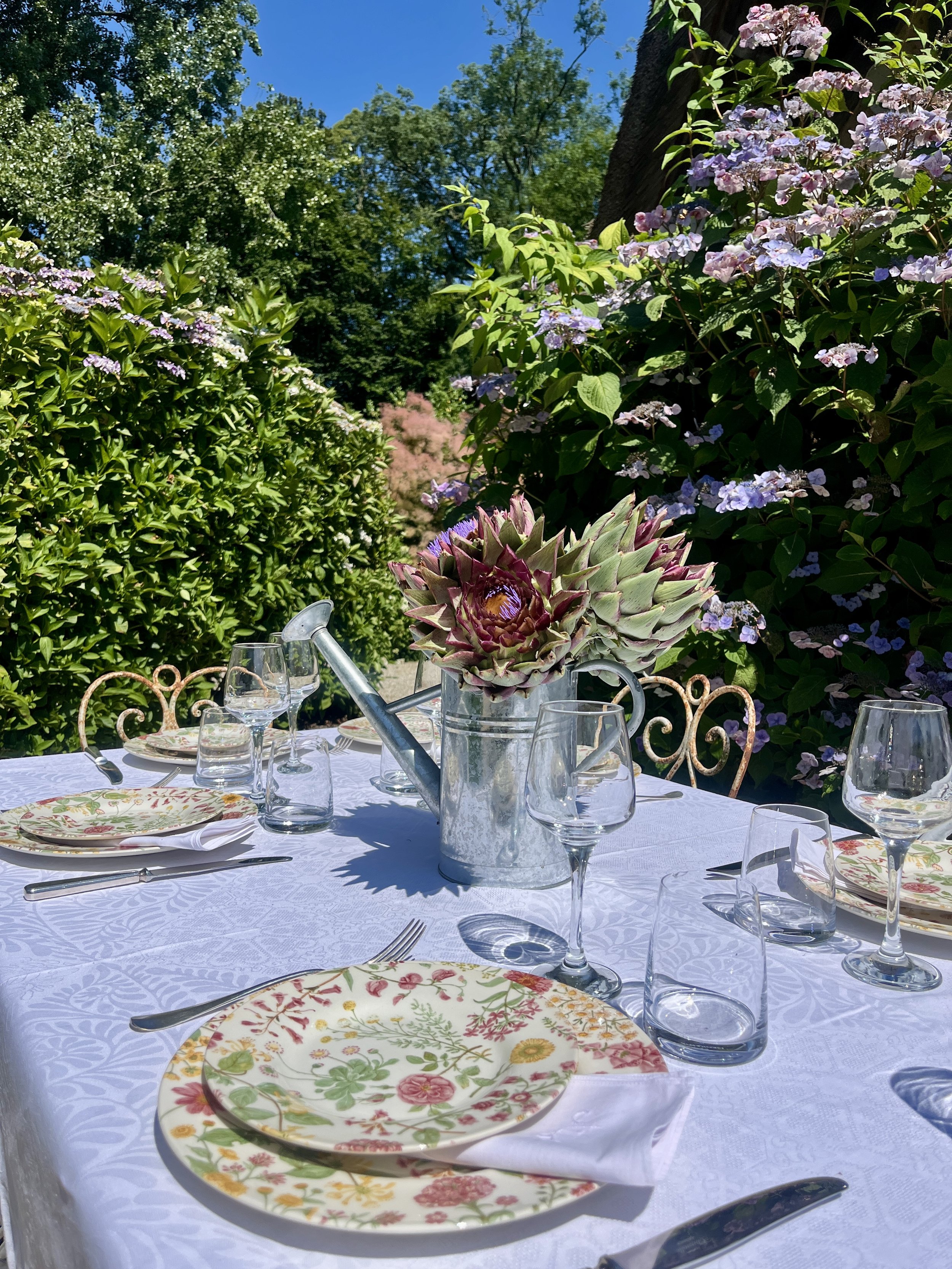 Une table extérieure élégante avec une nappe blanche, des assiettes à fleurs, des verres à vin, des couverts en argent, et un vase avec un artichaut comme centre de table. En arrière-plan, des buissons et des arbres sous un ciel bleu clair.