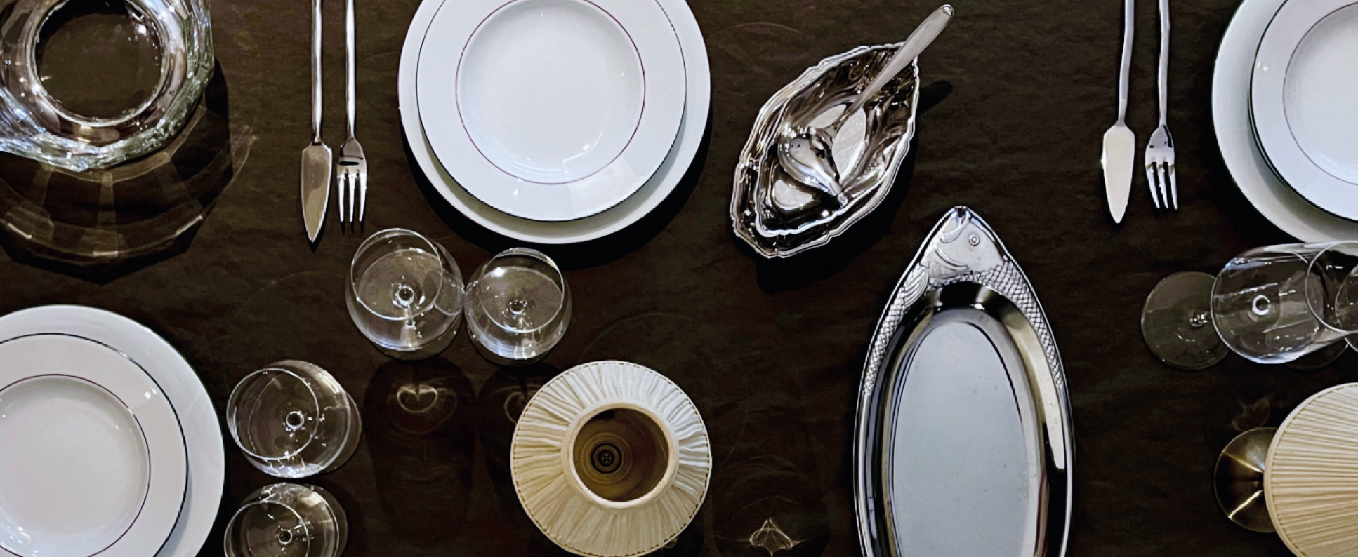 Table avec assiettes en porcelaine blanche, verres à vin, couverts en métal, plats en argent, et une bougie, préparée pour un dîner élégant.