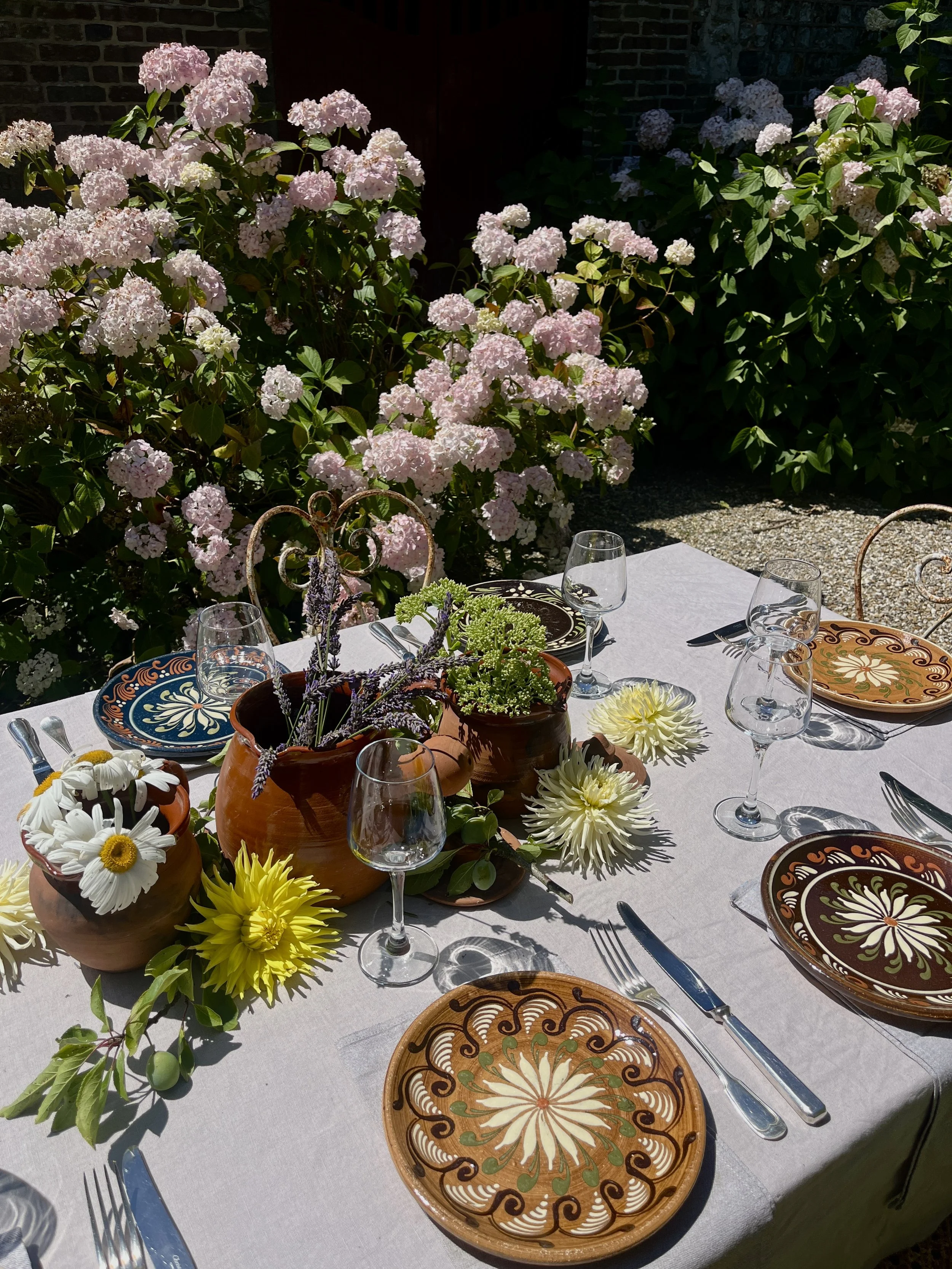 Table de repas en extérieur avec vaisselle en terre cuite décorative, fleurs colorées et purées, entourée de fleurs d'hortensias roses et de verdure.