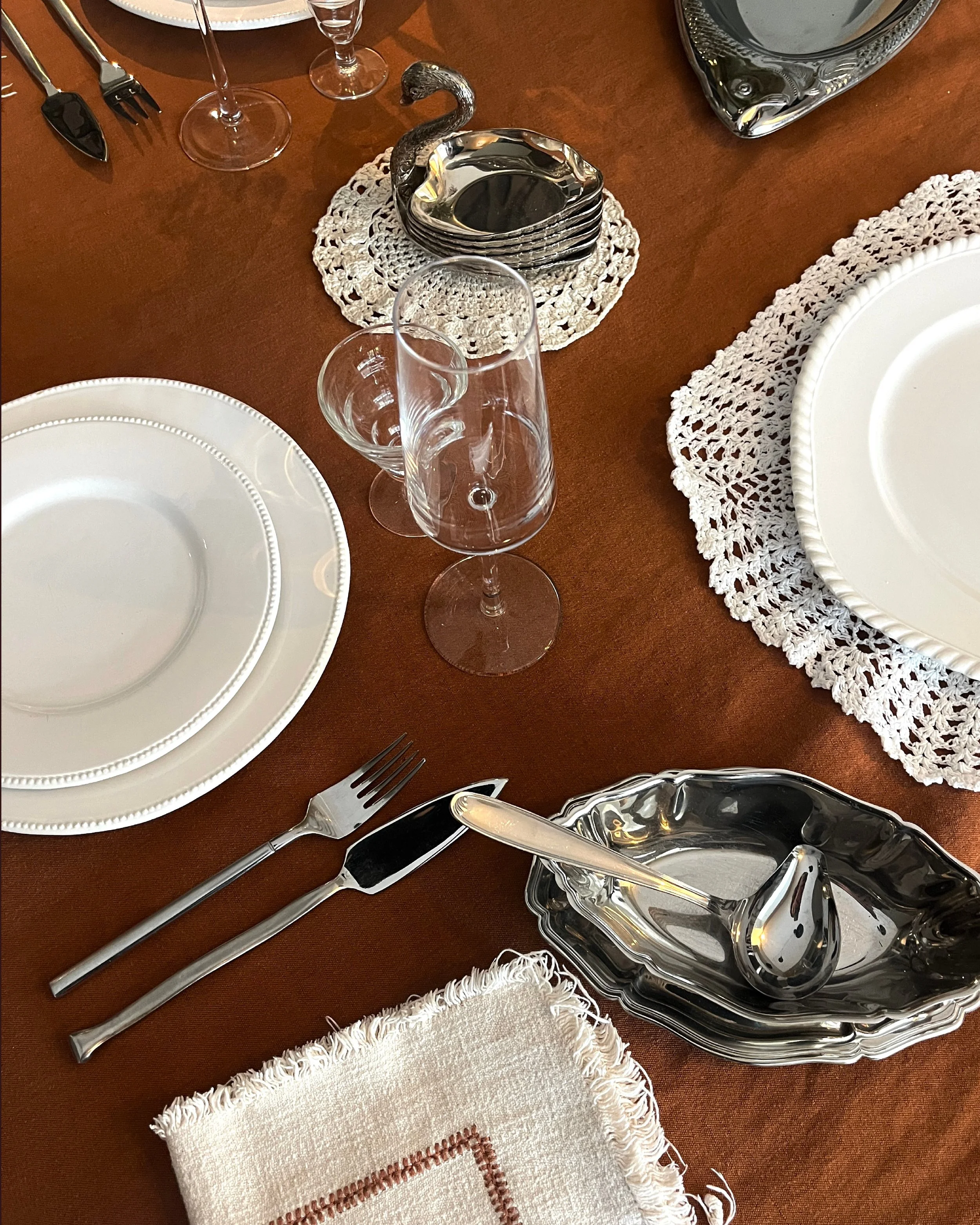 Une table élégante avec vaisselle blanche, couverts en métal, verres à vin, assiettes, nappe en tissu marron et sets de table en crochet blanc.