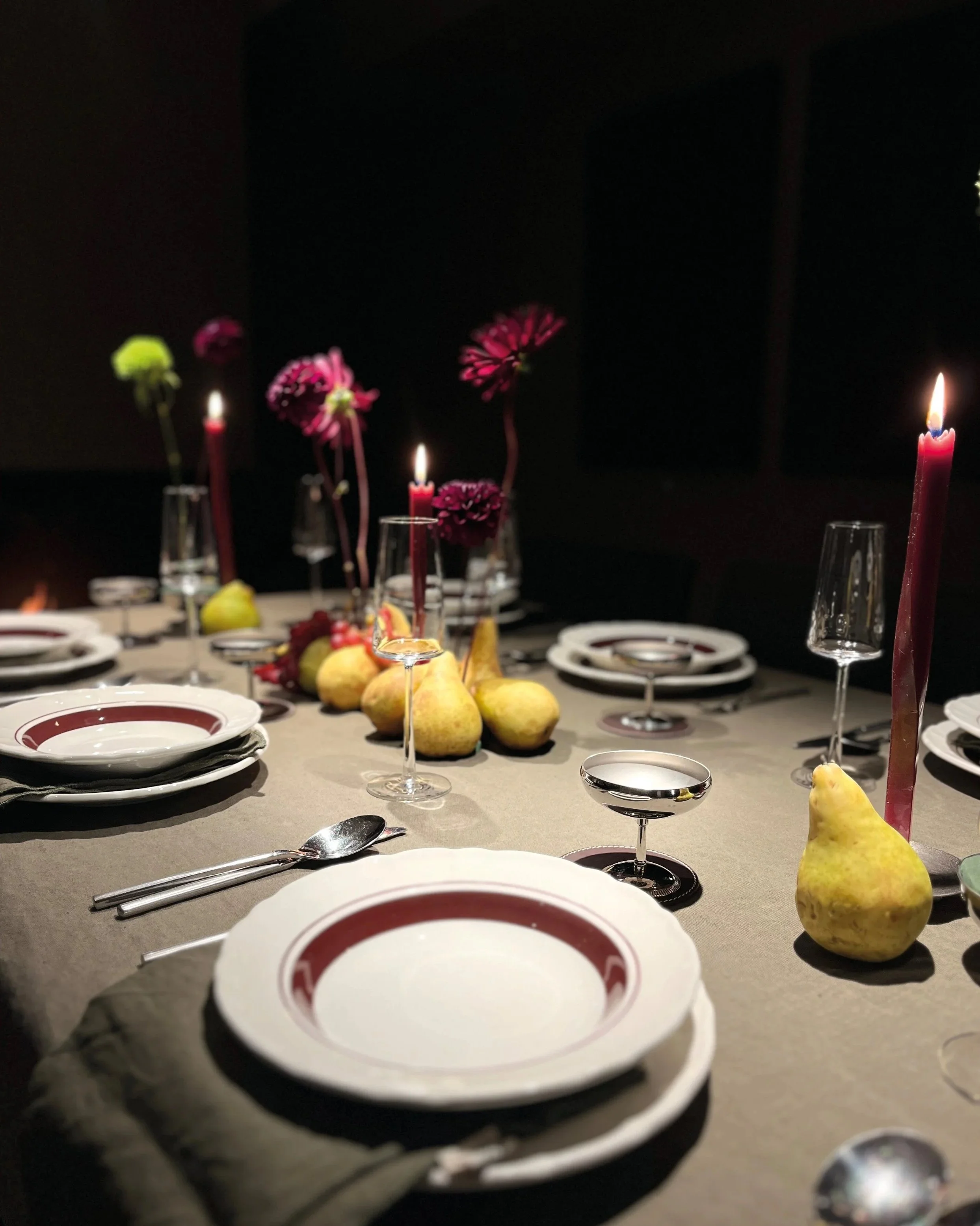 Une table élégante dressée pour un dîner, avec des assiettes, des couverts, des verres à vin, des chandelles allumées, et des poires en décoration, dans un cadre sombre et sophistiqué.