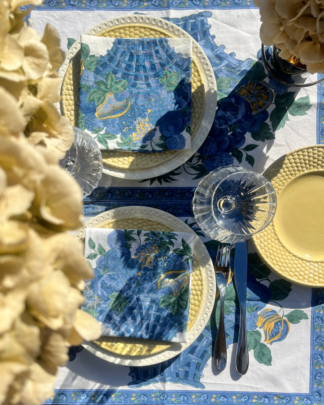 Une table dressée avec des assiettes, des verres, des couverts et des serviettes à motifs floraux bleus et verts, avec des décorations de fleurs sur la nappe. Le tout dans un style élégant et estival.