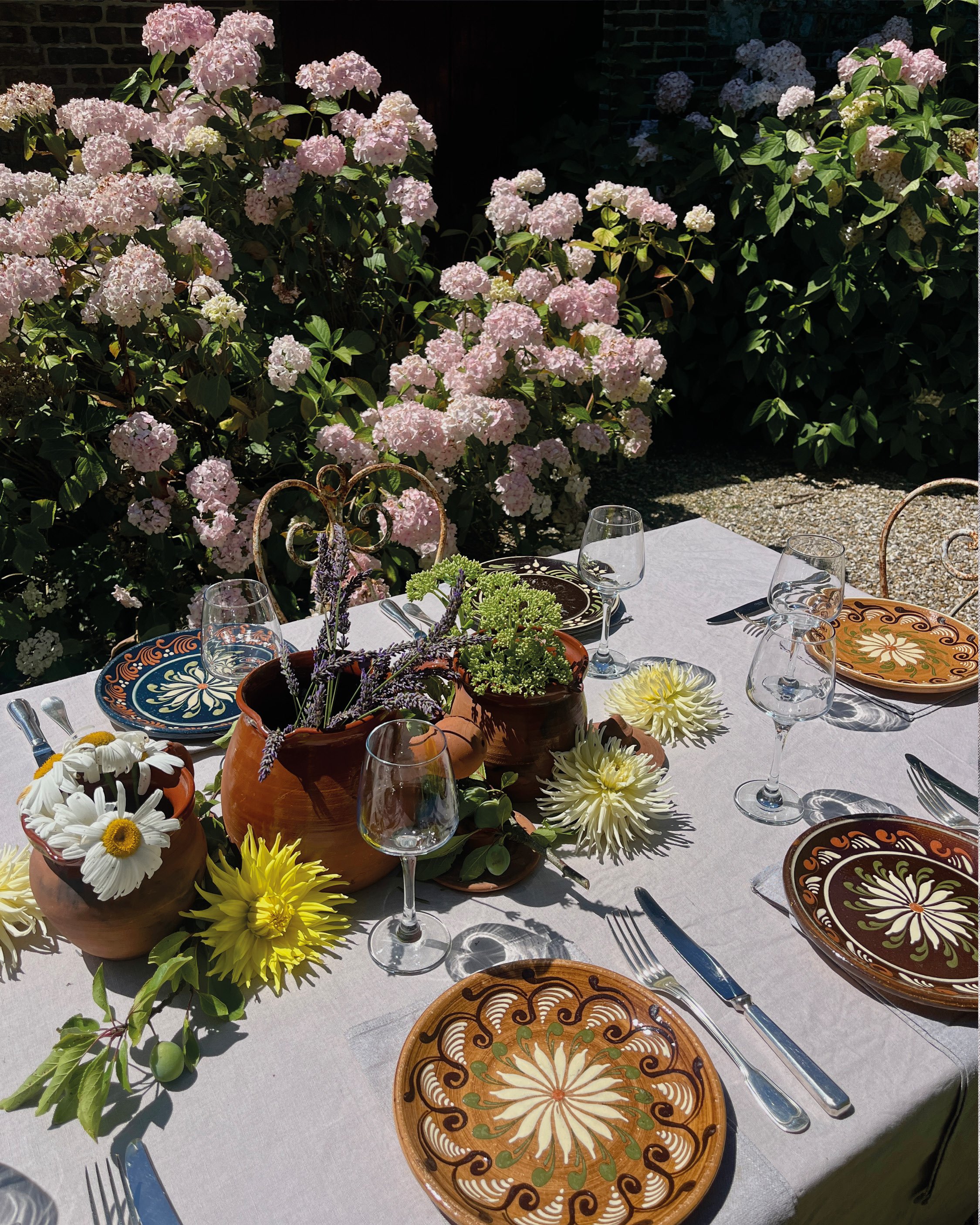 Table de repas en extérieur avec vaisselle en céramique décorative, verres à vin, et un centre de table avec des fleurs, entourée de buissons de hortensias roses.