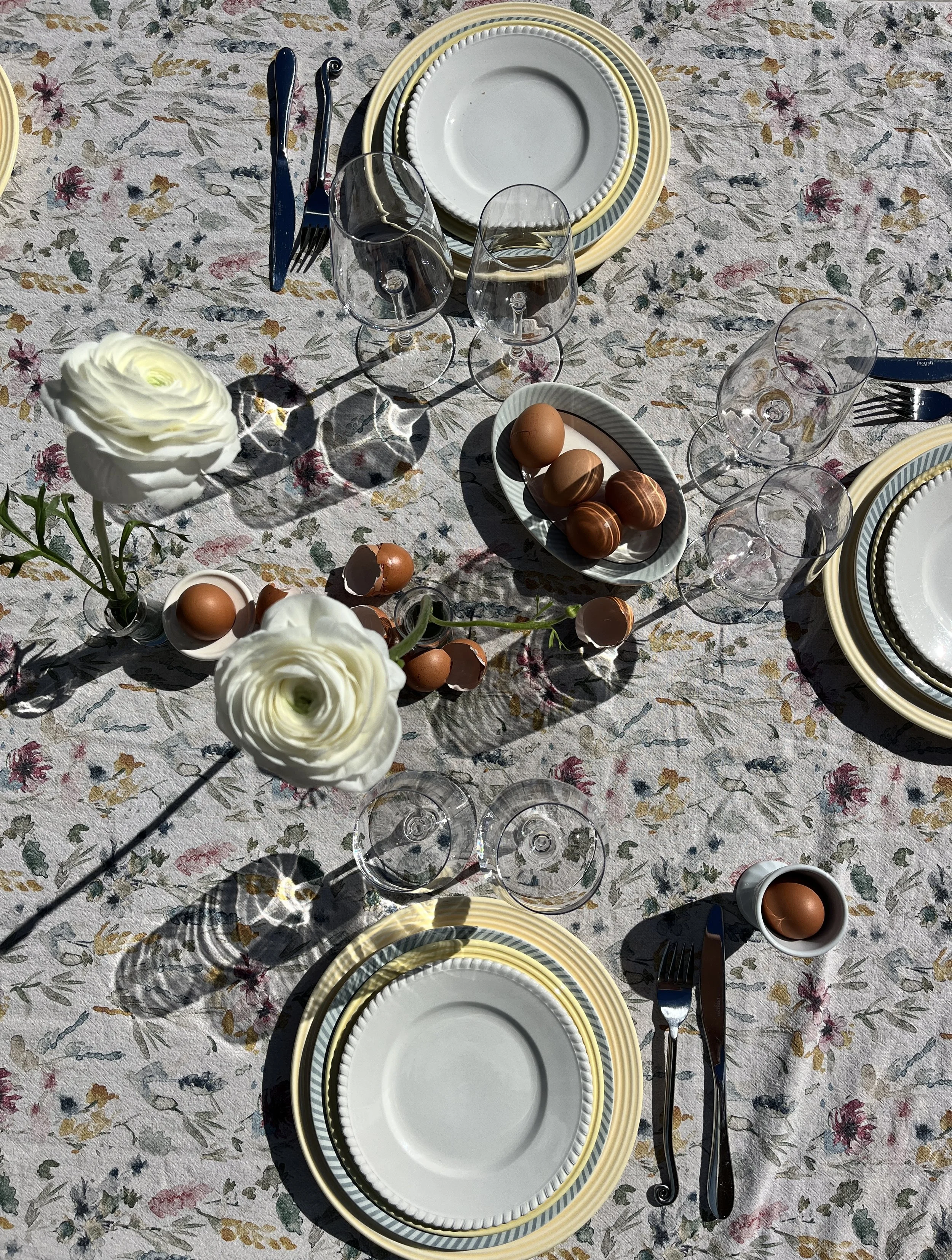 Table dressée pour un repas en plein air avec des assiettes, des couverts, des verres à vin, des œufs et des œufs en chocolat, décorée de fleurs blanches et de branches avec des œufs en chocolat.