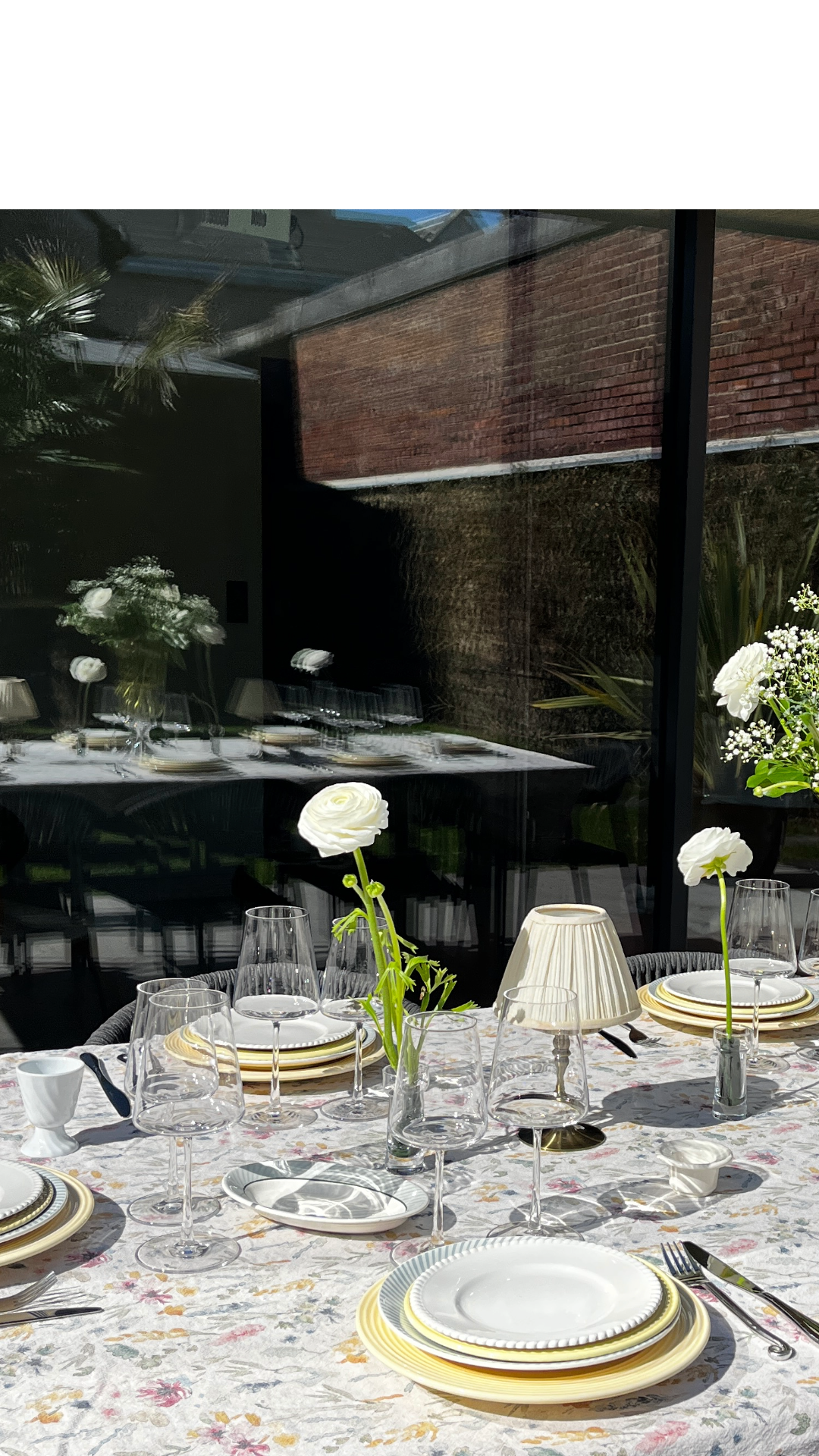 Table dressée avec de la vaisselle, des verres, des fleurs blanches, une lampe de table et une nappe florale, vue à travers une vitre reflectante.
