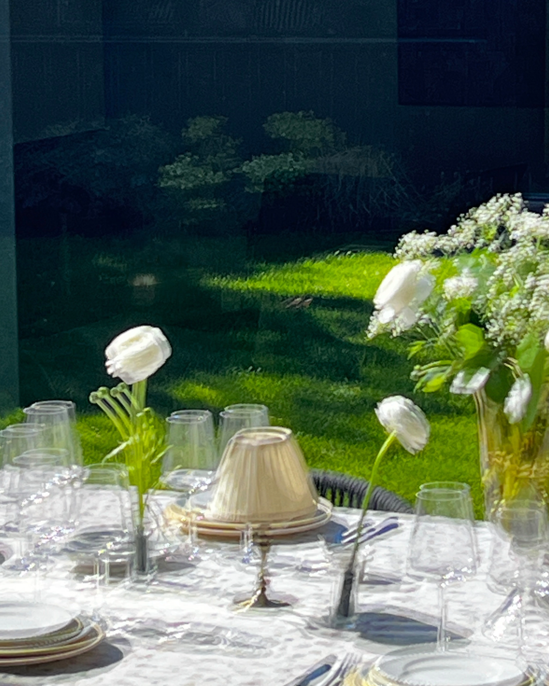 Image floutée laissant deviner Table dressée avec des verres, des assiettes, des fleurs blanches dans des vases, et une lampe en laiton, avec un jardin en arrière-plan.