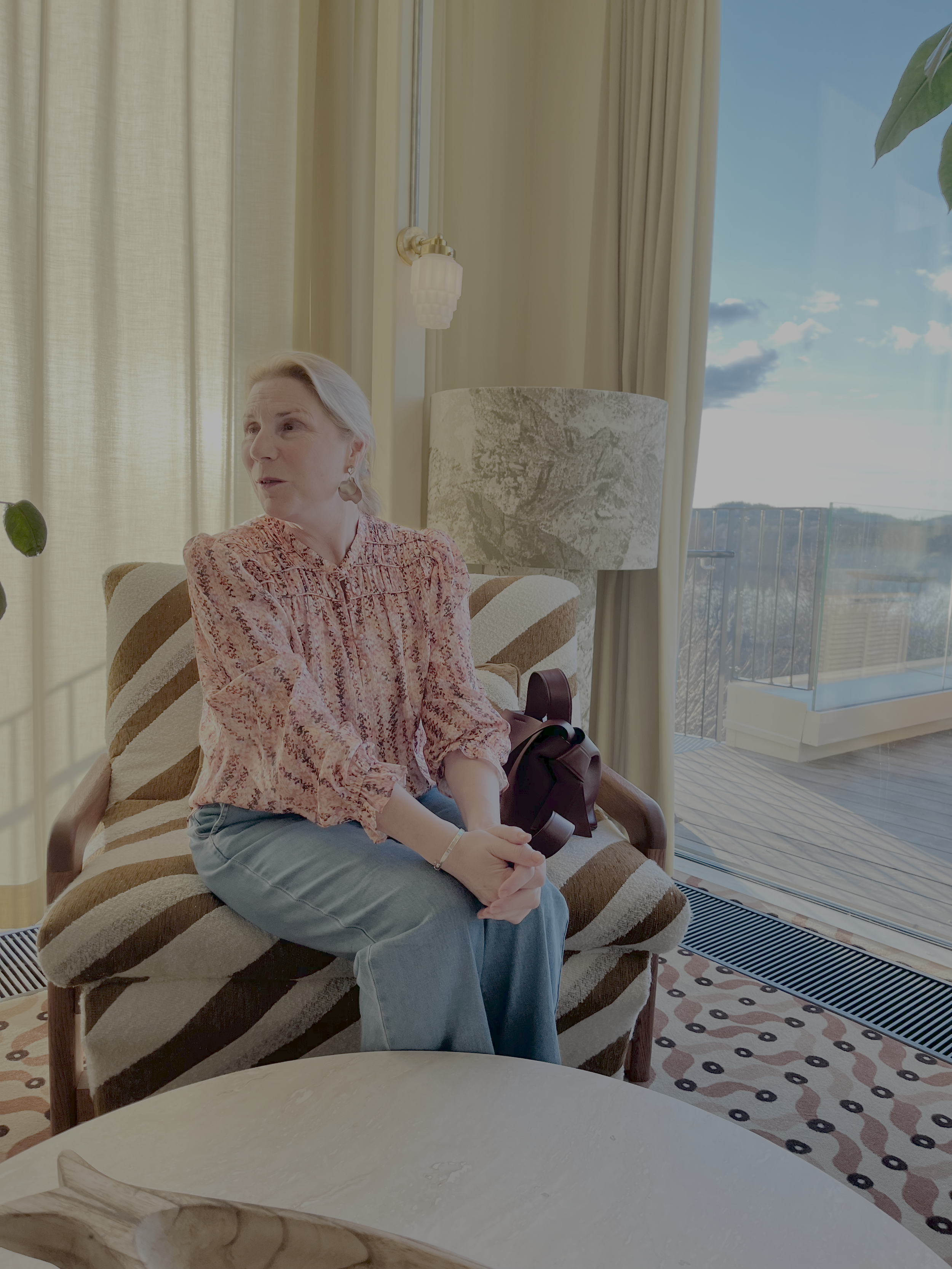 An older woman with light hair sits on a patterned armchair in a brightly lit room, holding a black object in her hands, with a large window and a balcony outside in the background.