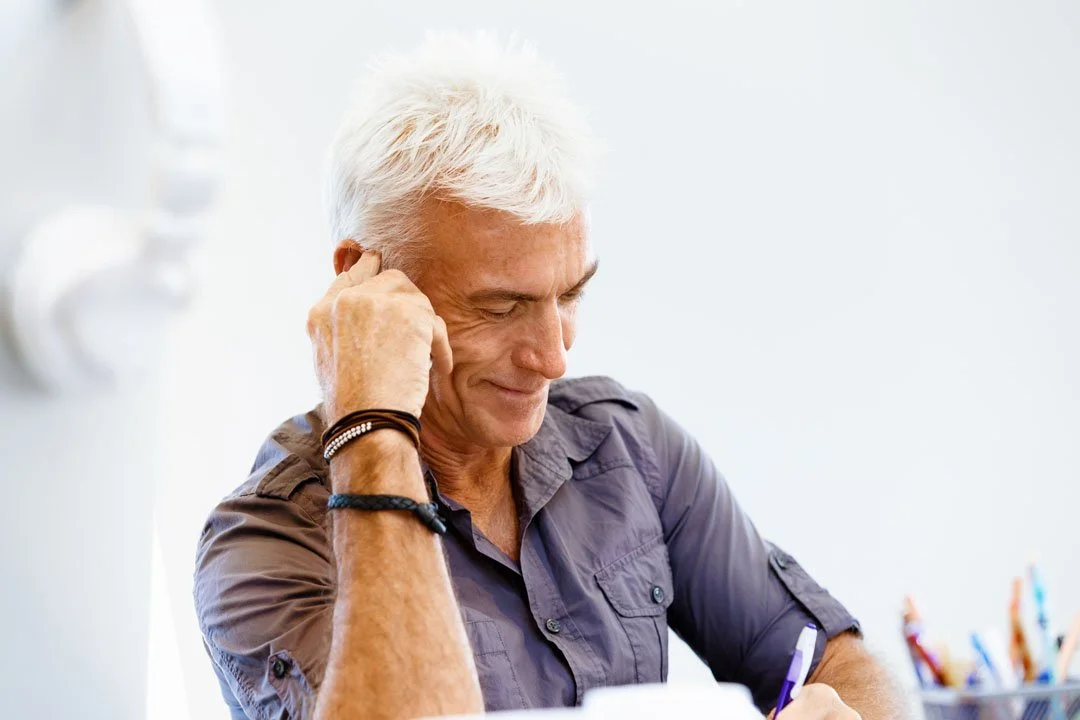 Foto eines älteren Mannes mit weißen Haaren, aber noch gut in Form. Er stützt seinen Kopf auf seiner Faust ab und lächelt