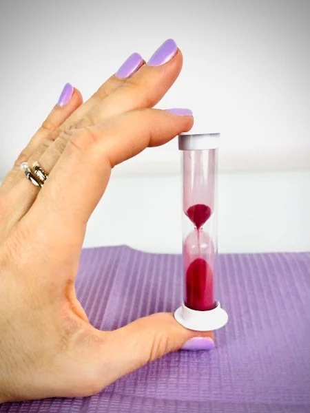 Eine Hand mit lilafarbenem Nagellack hält eine Sanduhr mit roten Sandkörnern über einer violetten Yoga-Matte.