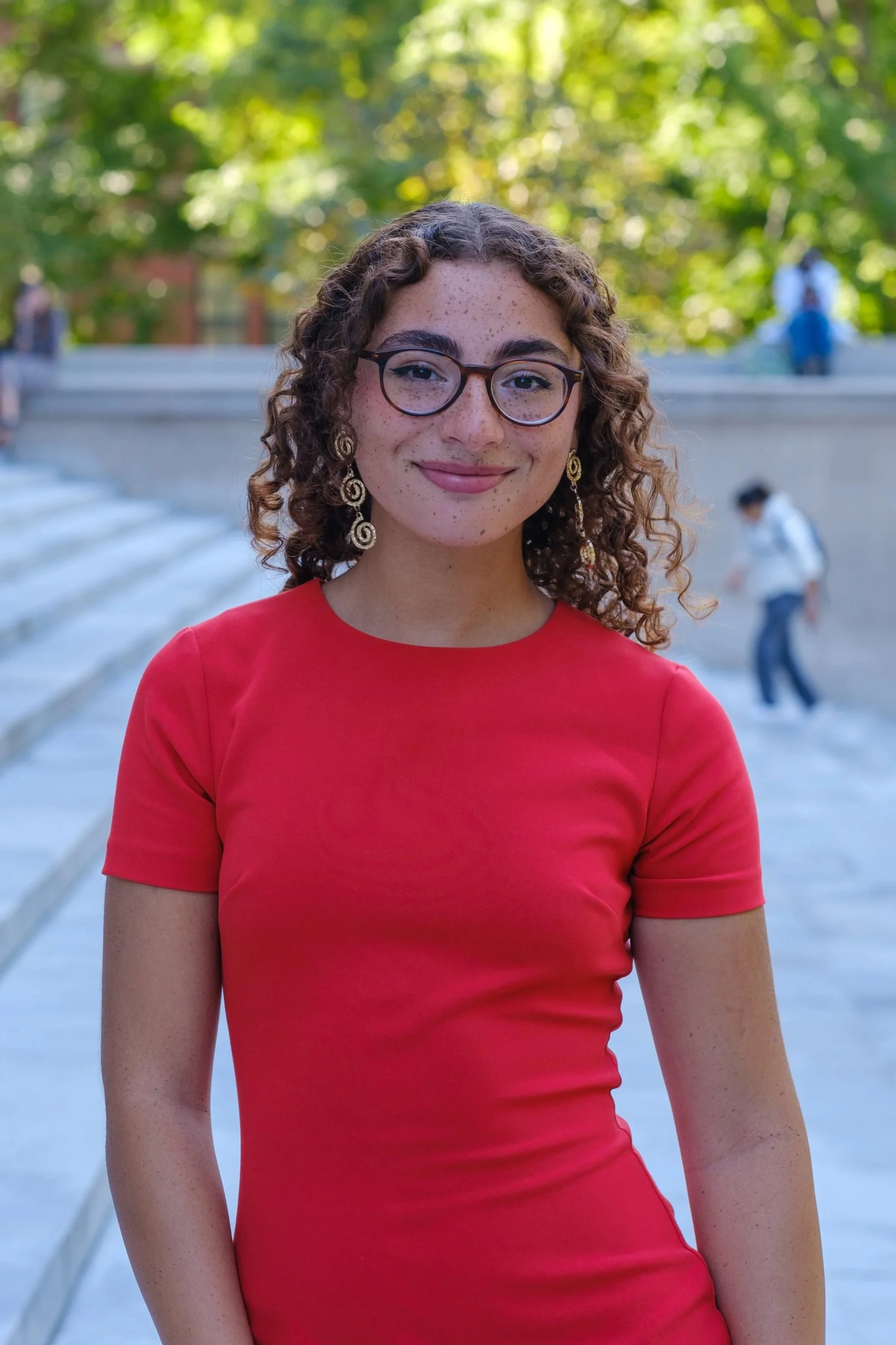 A smiling man wearing glasses, a white shirt, and a dark suit jacket stands indoors near a large wooden table, with blurred people and restaurant decor in the background.
