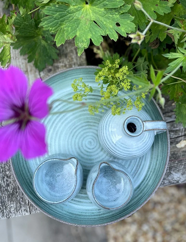 A green ceramic water fountain with a teapot-shaped spout and concentric circle design, surrounded by green leaves and purple flowers, on a weathered wooden surface.