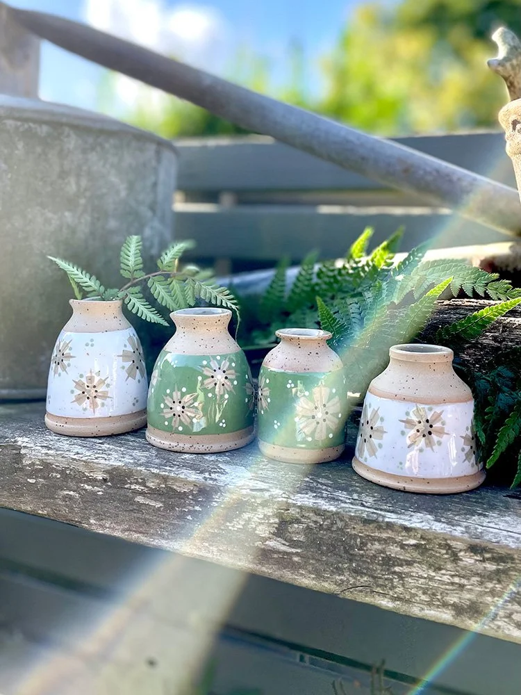 Four small ceramic vases with floral designs are placed on a weathered wooden surface outdoors, with green fern leaves and a stone planter in the background, illuminated by natural sunlight.