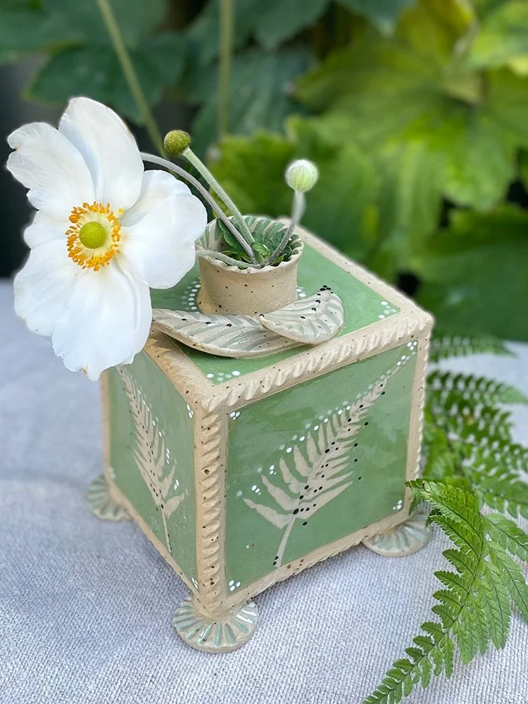 Decorative ceramic pot with a white flower and budding plant, set on a green and cream-colored stand with leaf patterns, surrounded by greenery.