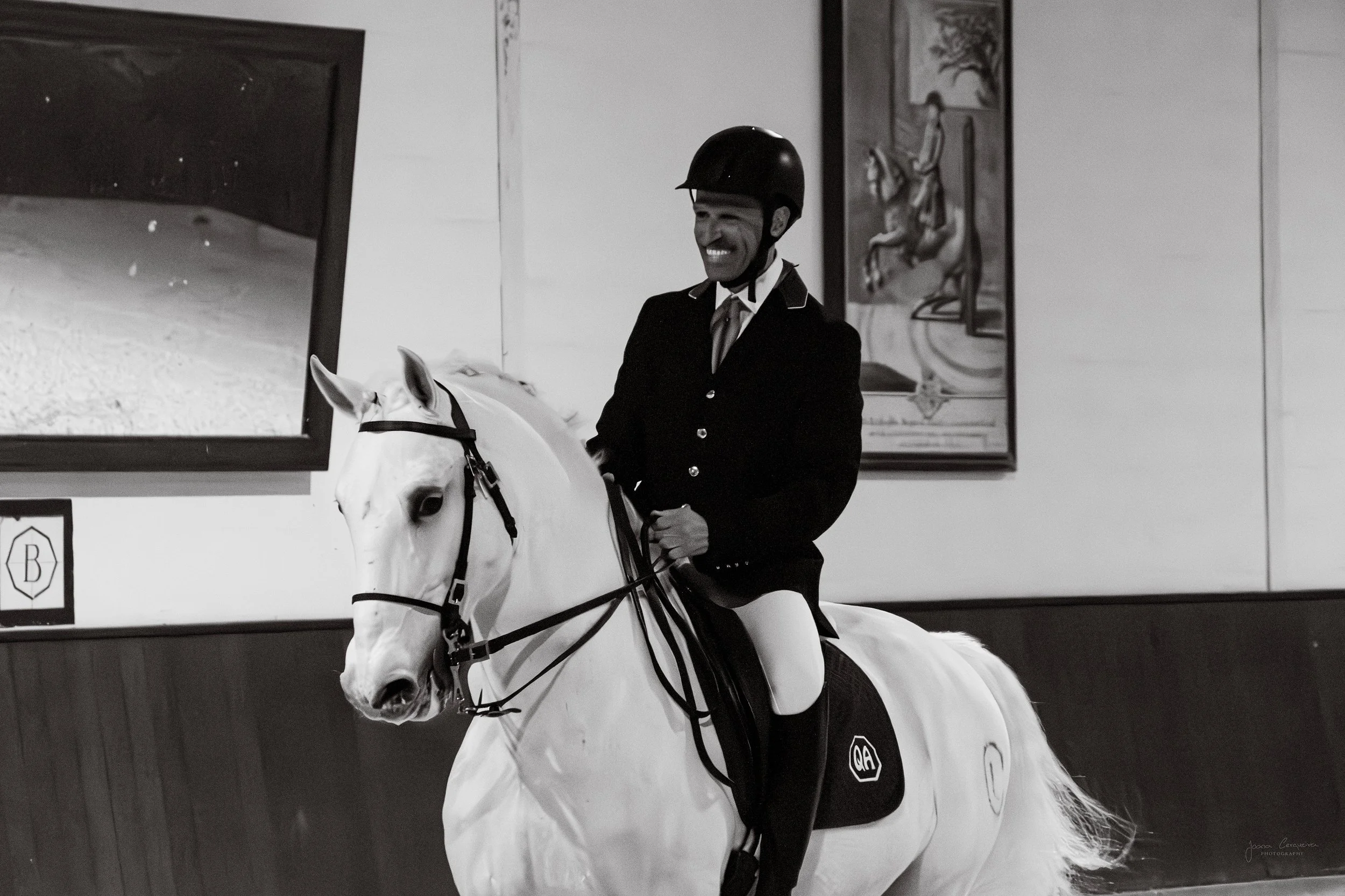 A man in a dark riding jacket, riding helmet, and white breeches sitting on a white horse indoors. The man is smiling.
