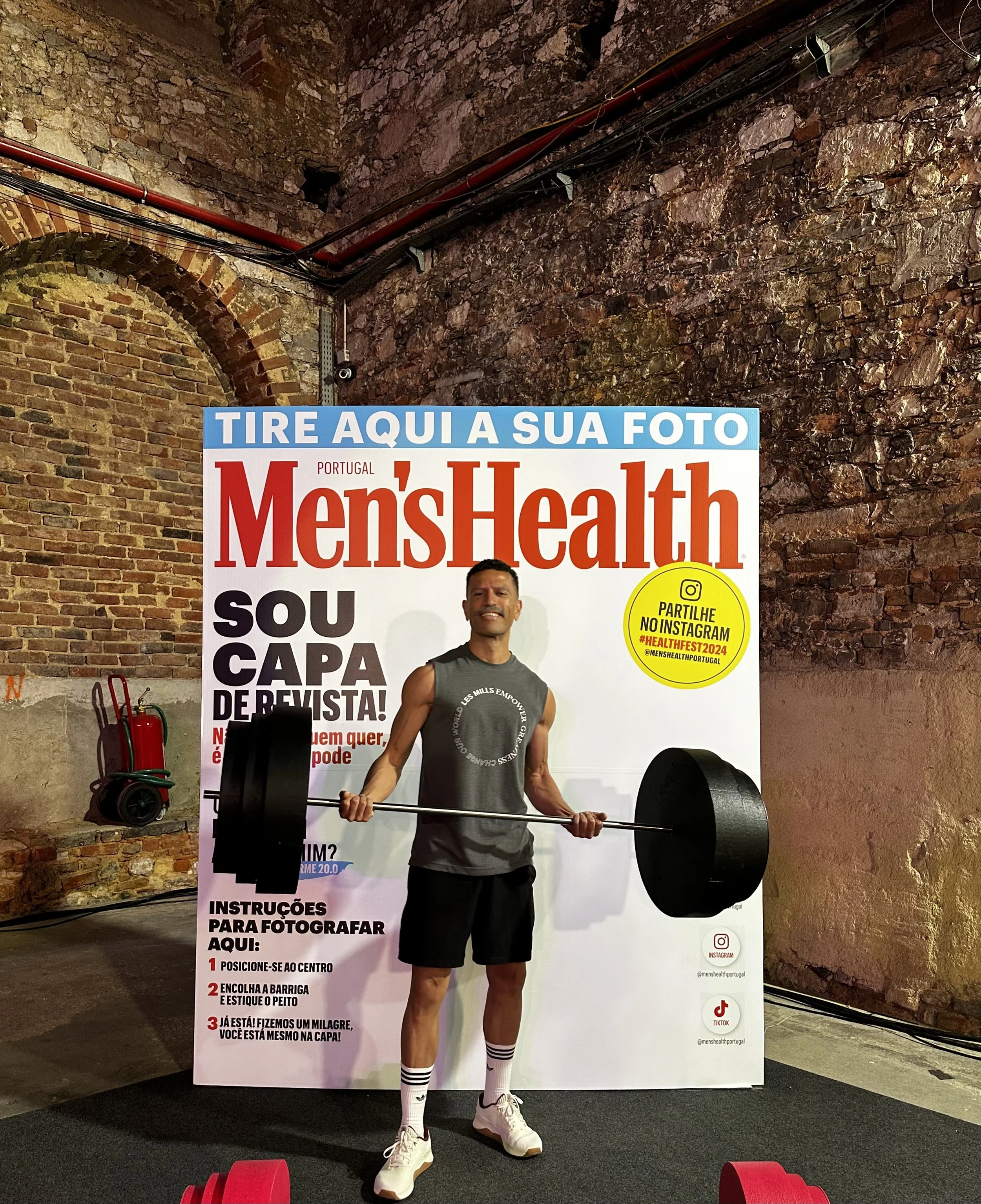 A man in athletic clothing holding a barbell with weight plates in front of a promotional display for Men's Health magazine. The background shows a brick wall and a fire extinguisher.