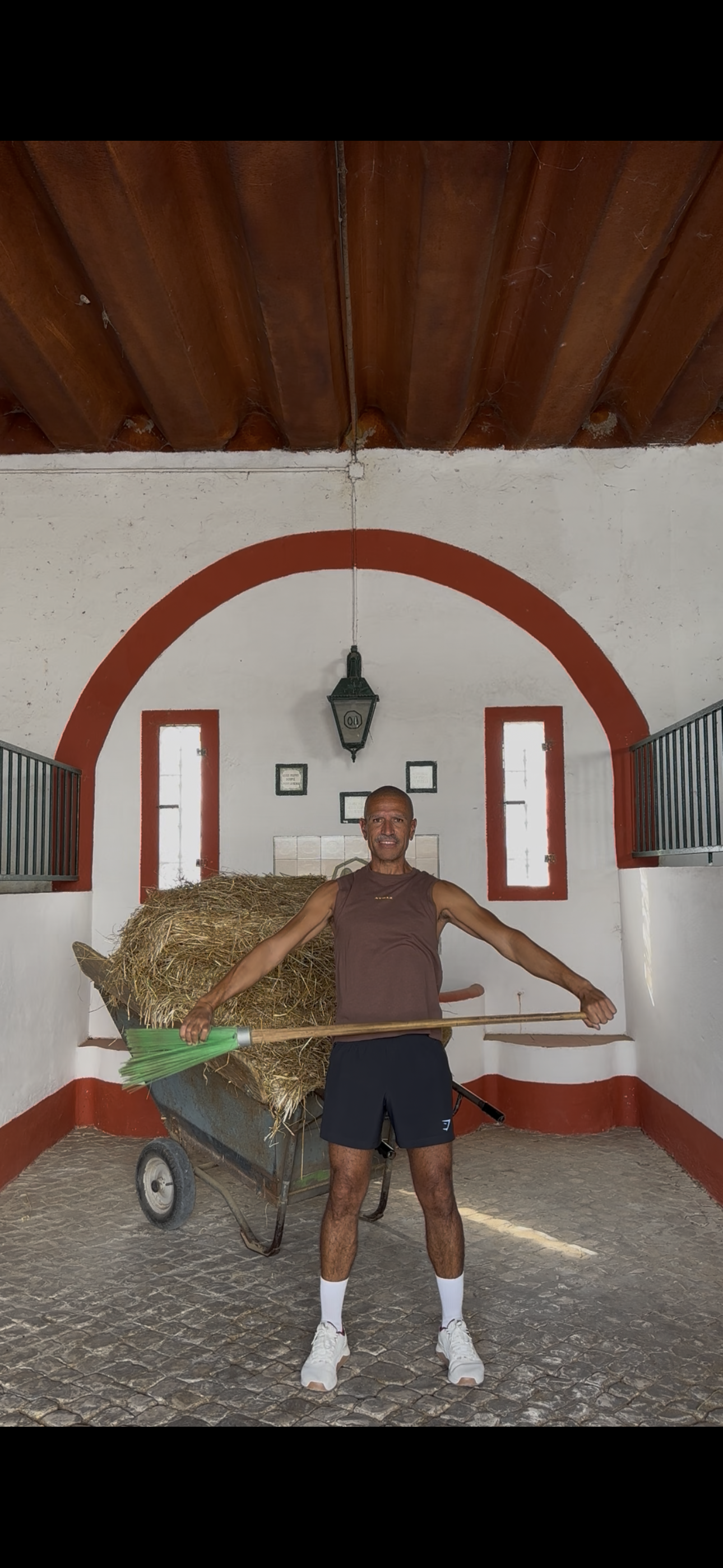 Man standing inside a room with a cart loaded with hay, holding a long broom, with decorative windows and a hanging lantern in the background.